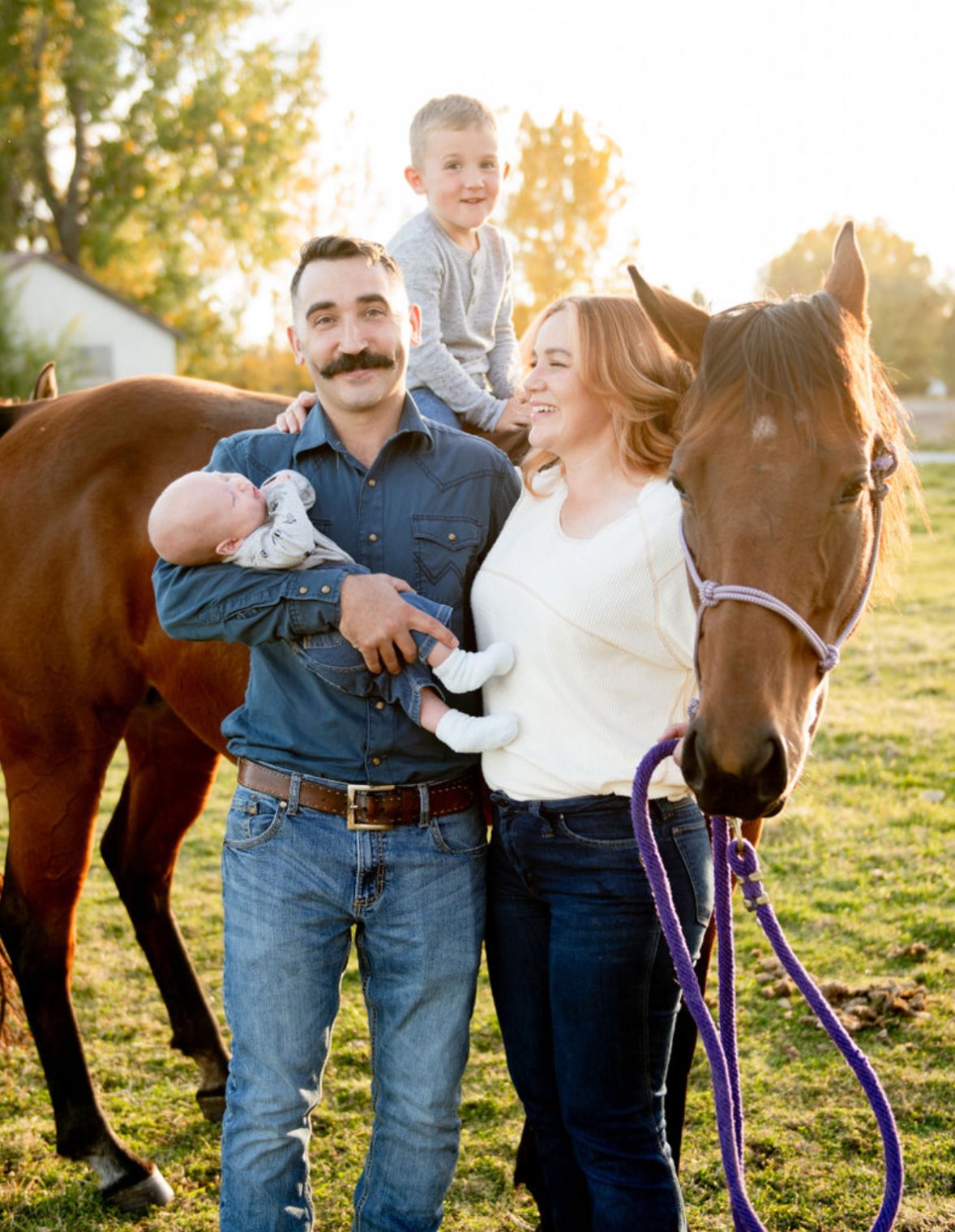 A family of four stands with a horse in a grassy park at sunset. The father holds a baby, the mother smiles at a young boy sitting on the horse, which is wearing a purple halter.