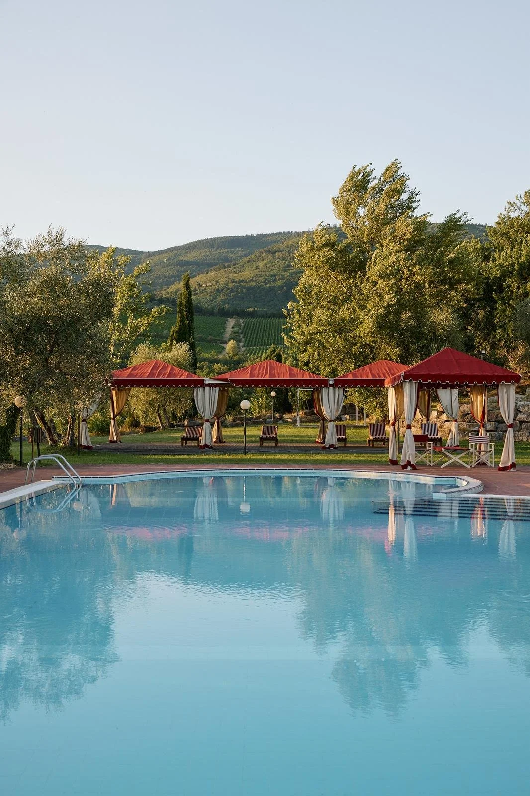 A swimming pool with reflections, surrounded by trees and red canopies with curtains and lounge chairs, with green hills and a clear sky in the background.