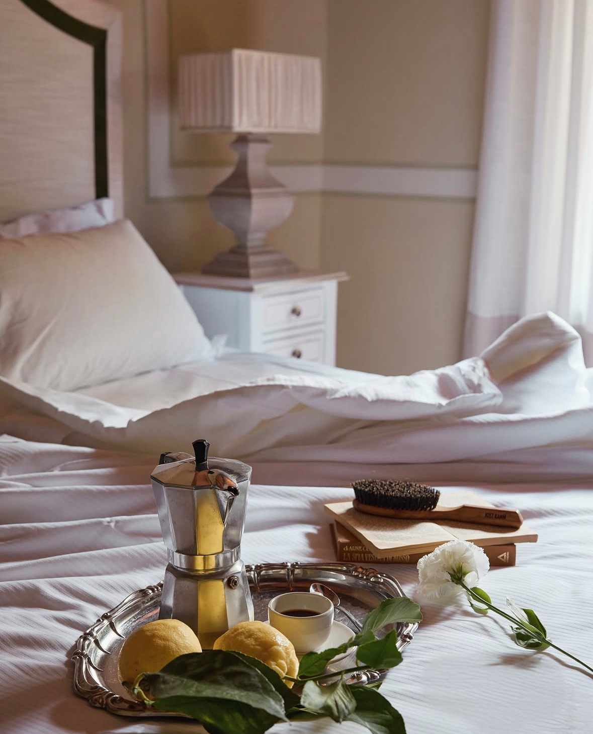 A cozy hotel bed setup with a silver tray holding a moka pot, lemons, a small cup of coffee, and greenery; a white pillow, a wooden book, and a hairbrush are on the bed; a bedside table with a lamp and beige curtains are in the background.