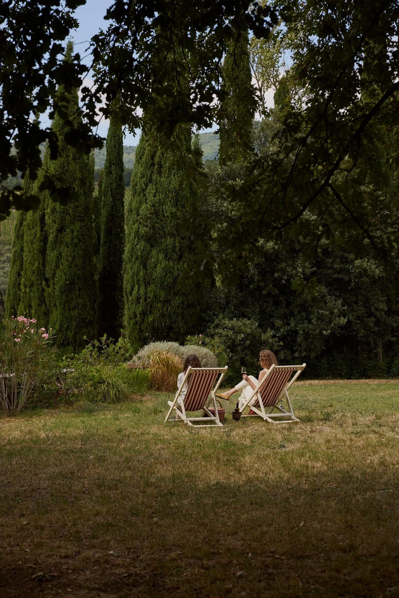 Two women sitting on deck chairs in a garden, drinking wine, with a background of trees and a mountain, in the evening.