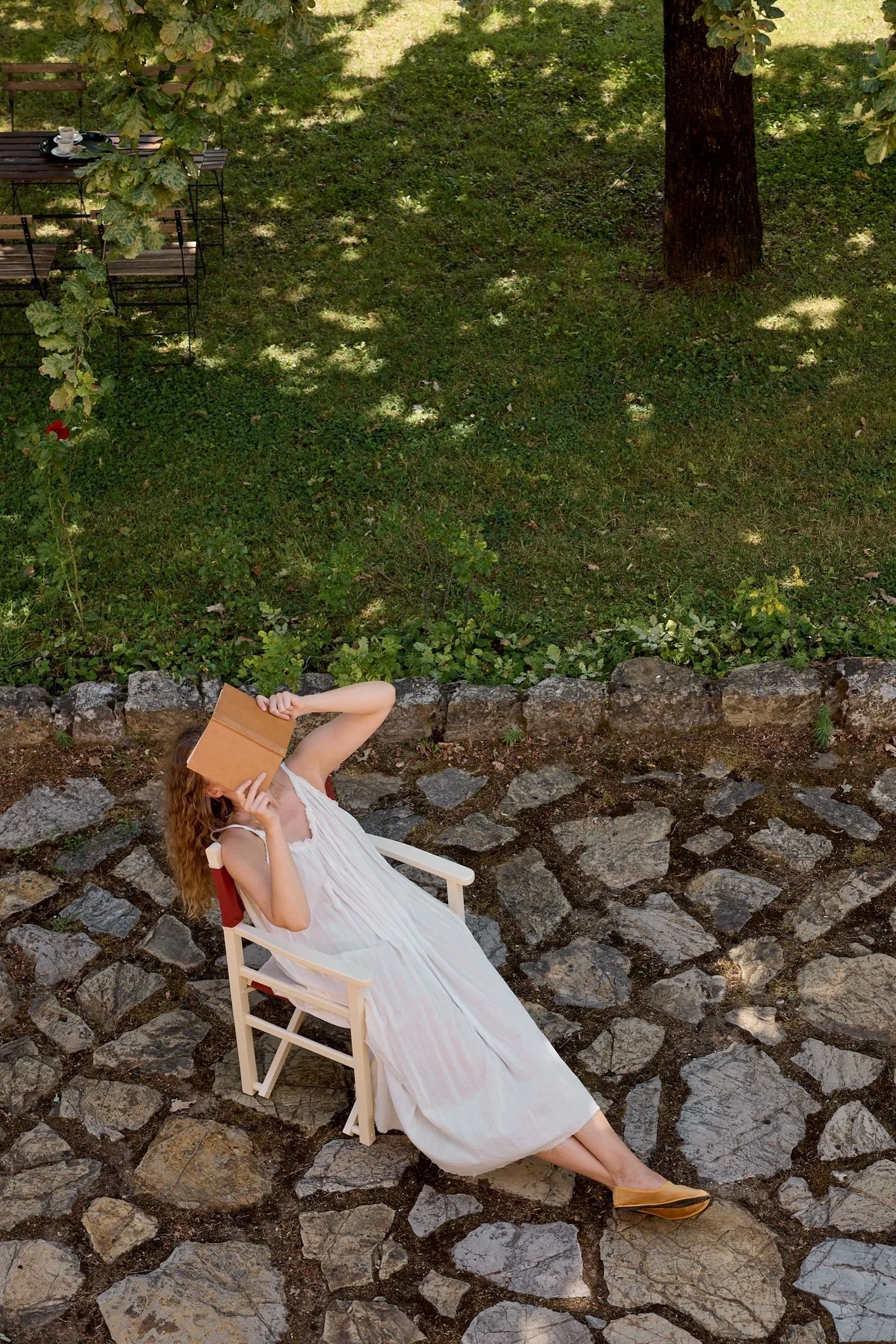 A woman with long curly hair is sitting outdoors on a white chair, holding a book to her face. She is wearing a white sleeveless dress and tan shoes. The background shows a stone patio, a grassy area, and a large tree with shadows on the ground.