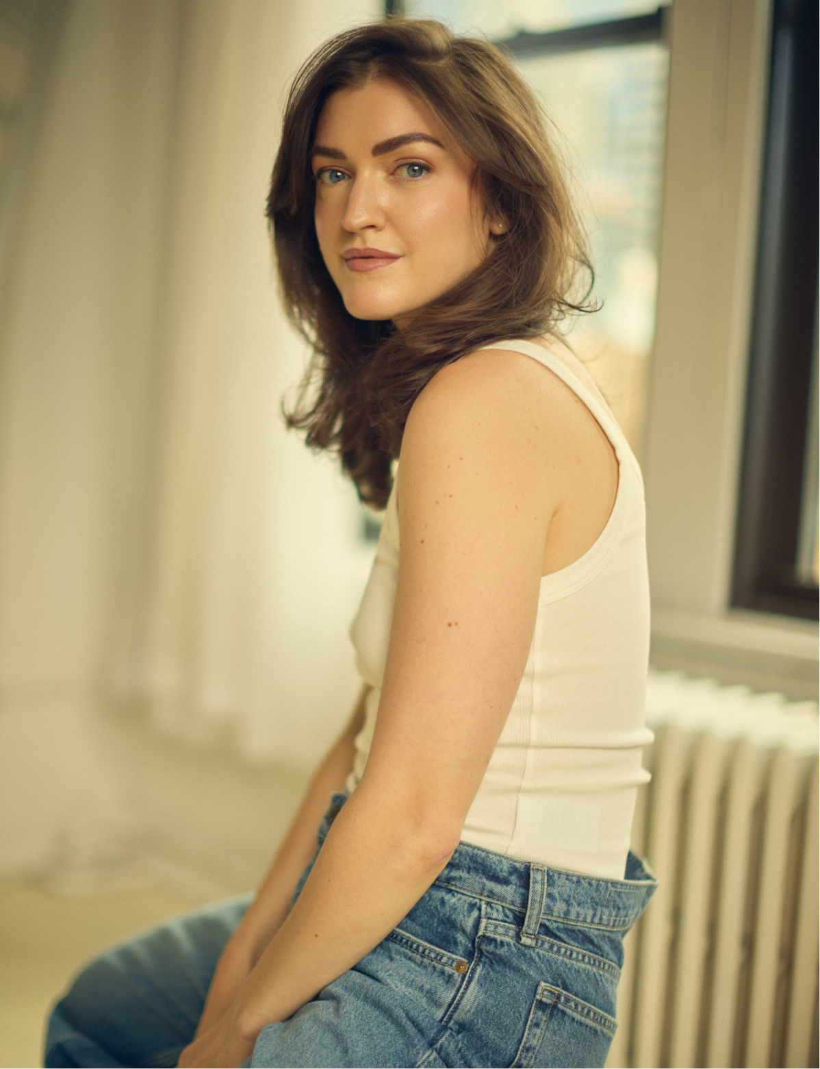 A young woman with brown hair and blue eyes sitting near a window, wearing a sleeveless white top and blue jeans, looking at the camera.