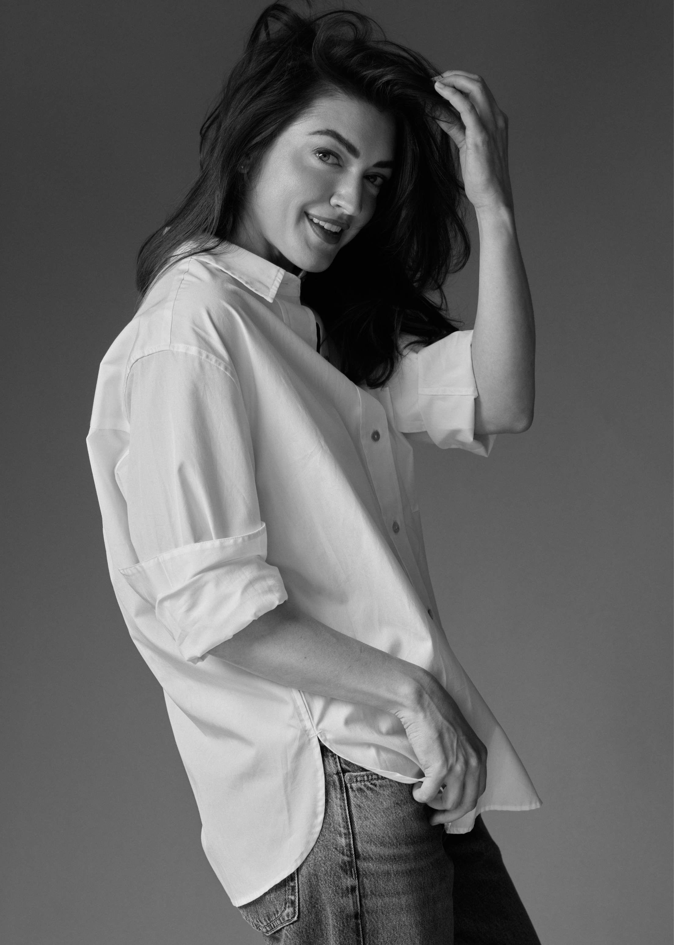 Black and white photo of a woman with long dark hair, wearing a loose fitting white shirt and jeans, smiling and touching her hair.