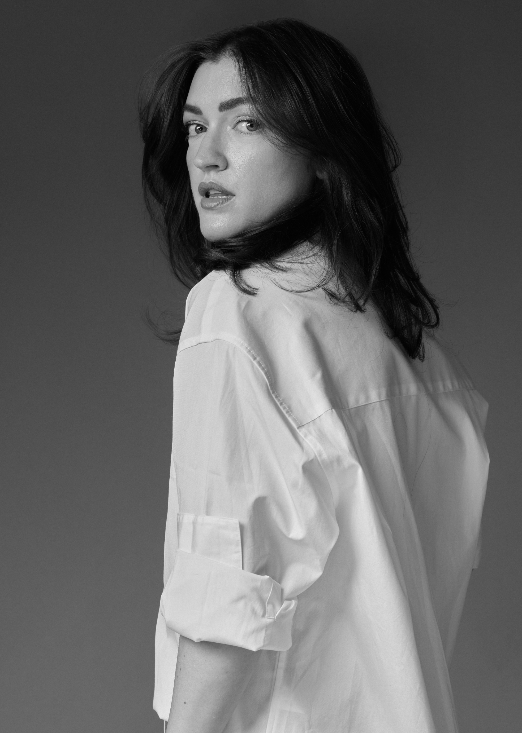 Black and white portrait of a woman with wavy hair, wearing a light-colored shirt, looking over her shoulder at the camera.