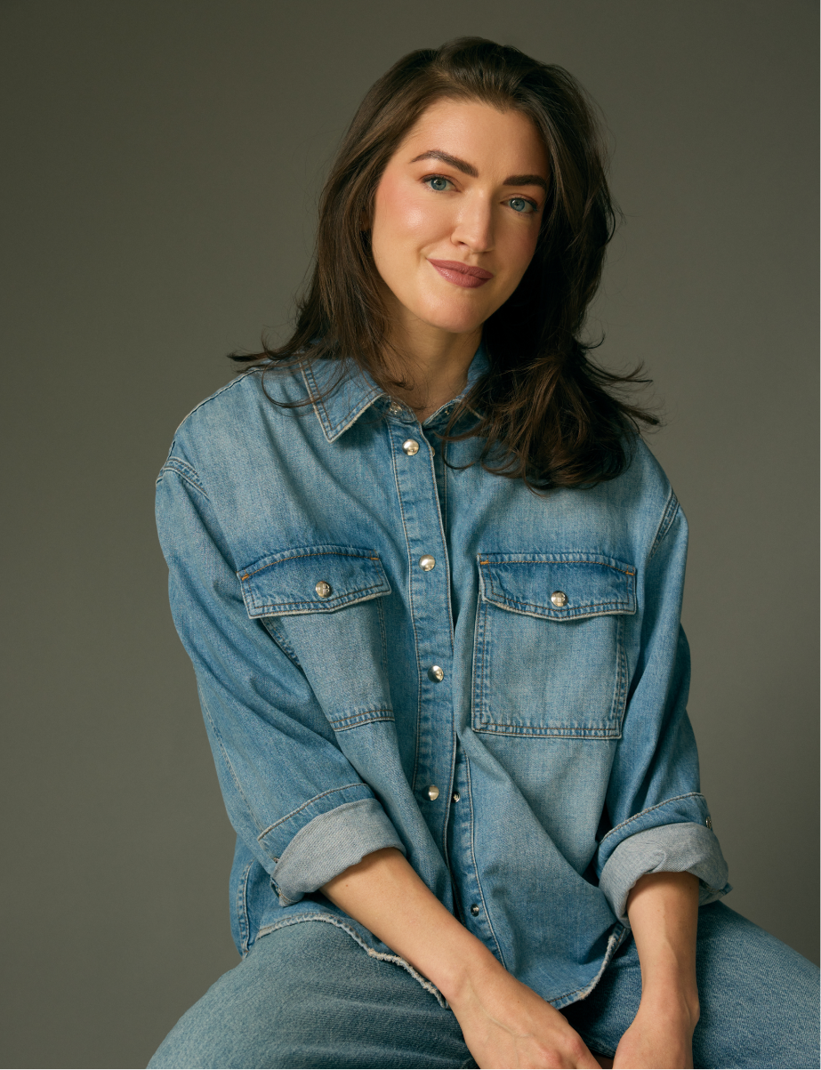 A young woman with brown hair and blue eyes wearing a denim jacket, sitting against a neutral background with a slight smile.