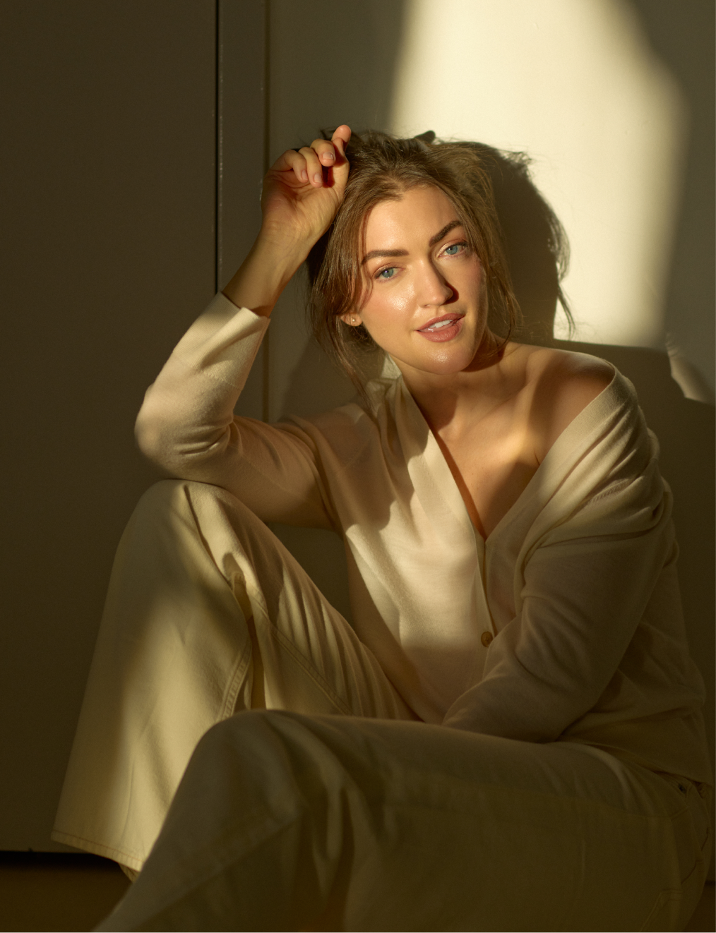 A woman with light skin, wavy brown hair, and blue eyes sitting on the floor, wearing light-colored pajamas, and posing in a warm sunlight showered room.