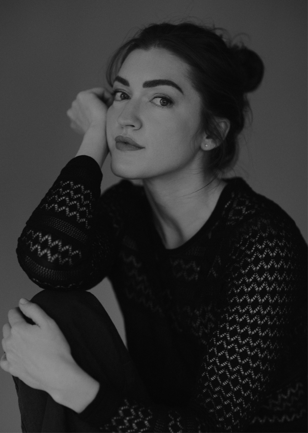 Black and white portrait of a young woman with dark hair in a bun, wearing a black lace top, sitting with her left arm resting on her knee, looking at the camera with a slight smile.