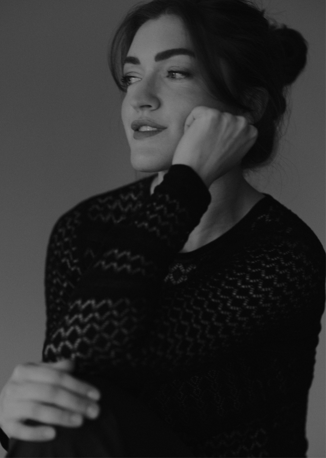 Black and white portrait of a woman with dark hair, wearing a lace long-sleeve top, resting her chin on her fist, looking thoughtfully to the side.