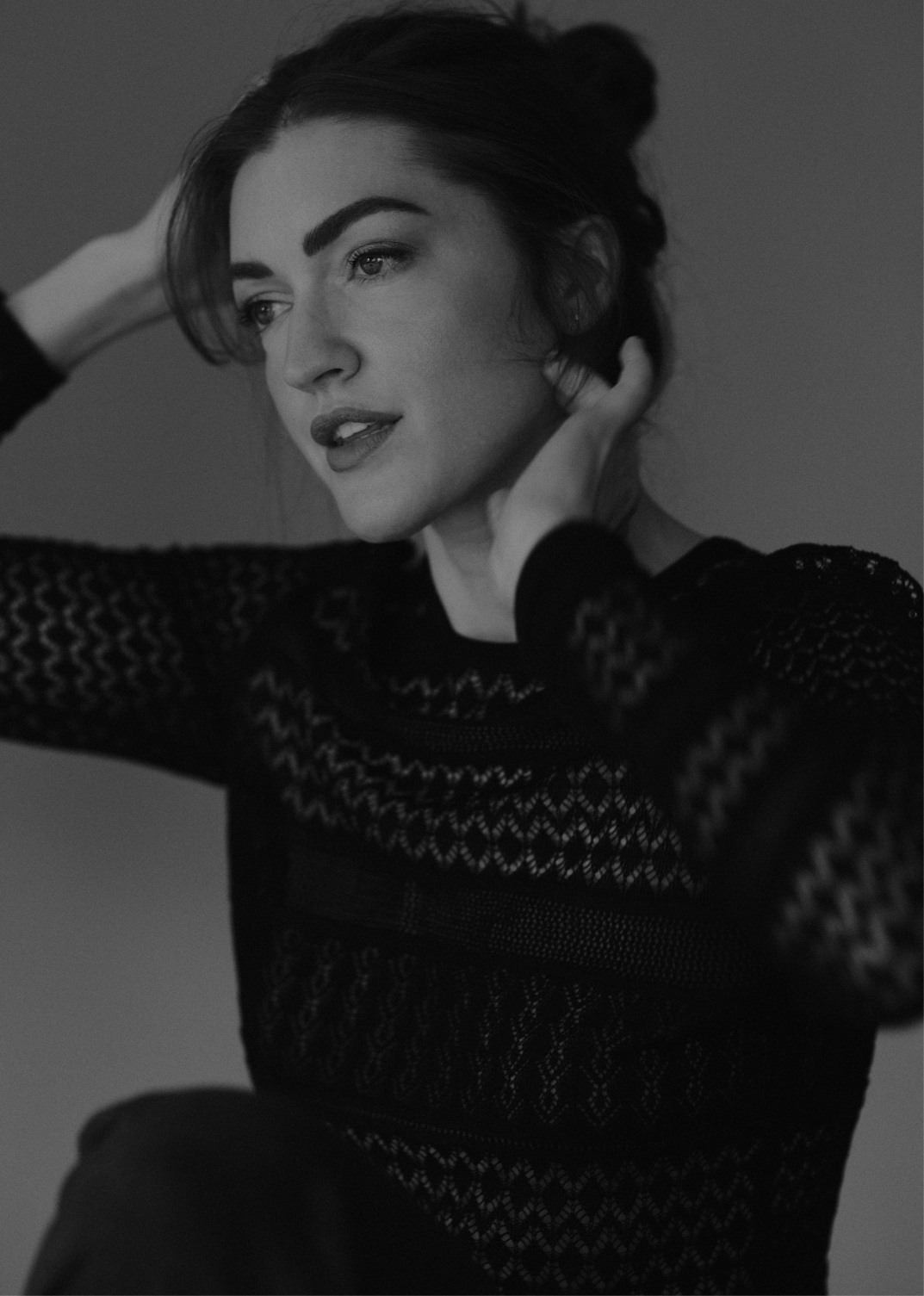 A black and white portrait of a woman with dark hair, wearing a lace patterned top, with her eyes looking to her left and her hands touching her hair and neck.