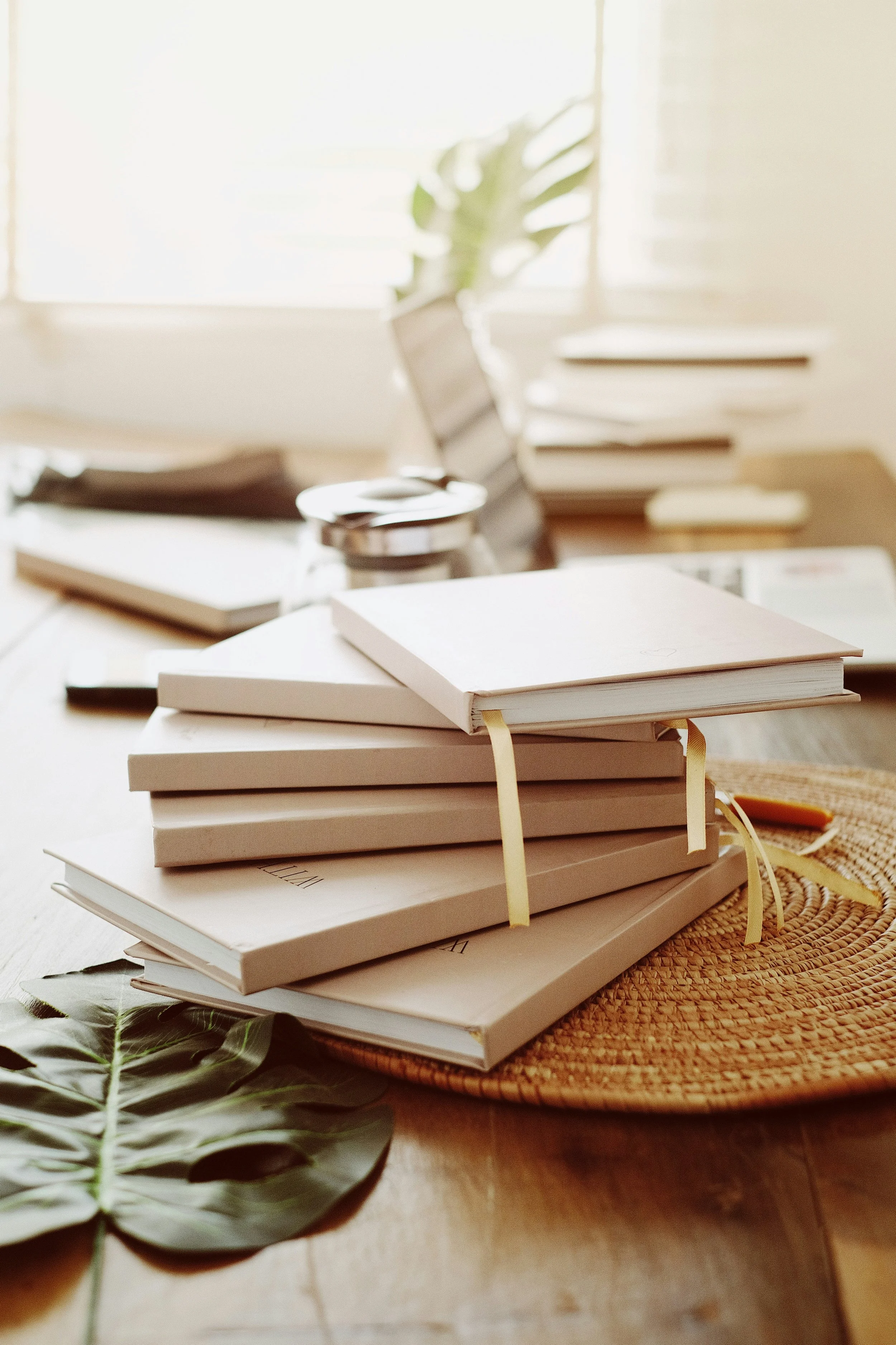 Stack of beige notebooks with yellowed ribbons on a wooden table, natural sunlight through a window, green leaf on table, woven placemat underneath.