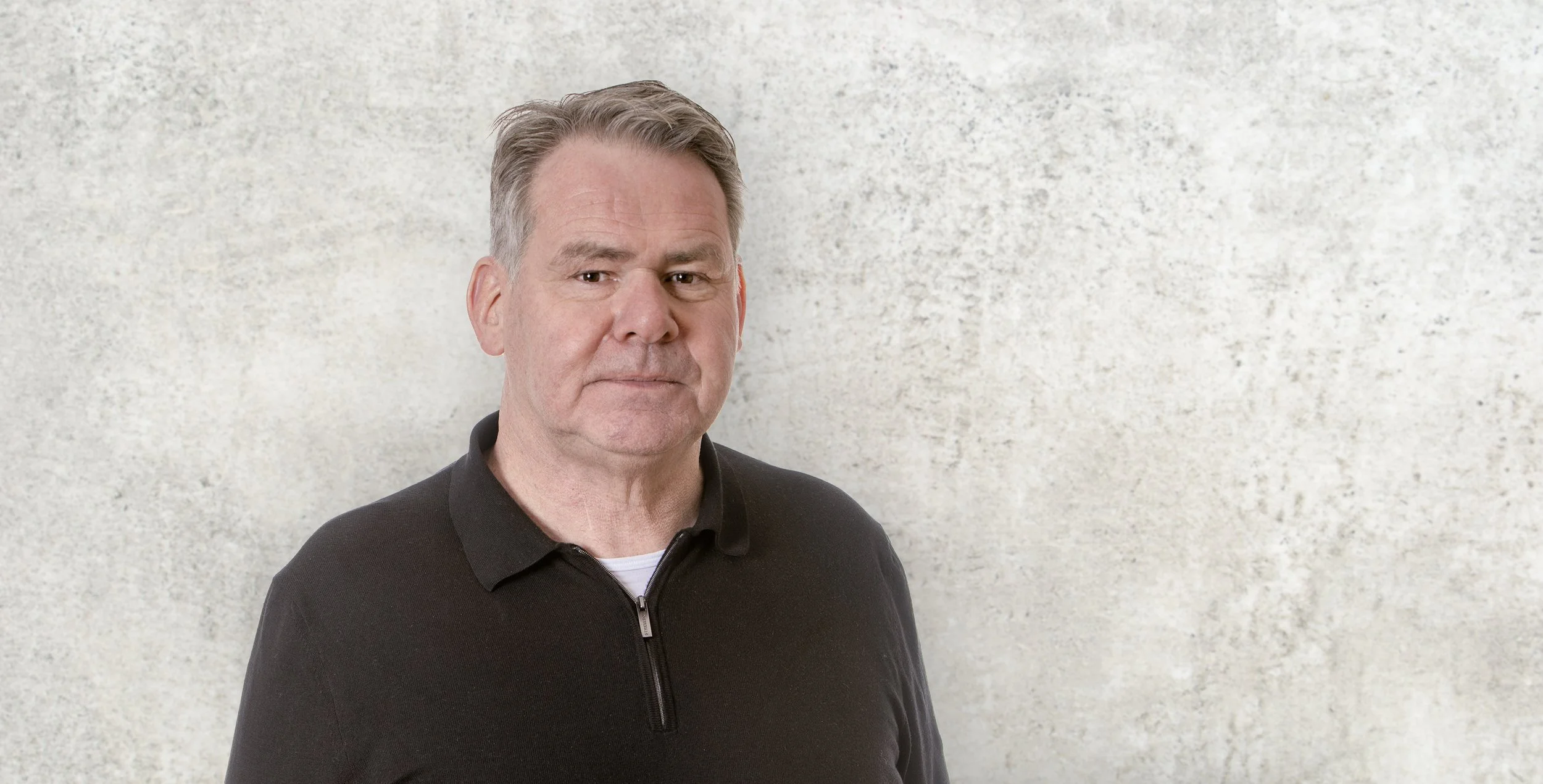 Ein Mann mit grauen Haaren, trägt ein schwarzes Polo-Shirt, steht vor einer hellgrauen Wand.