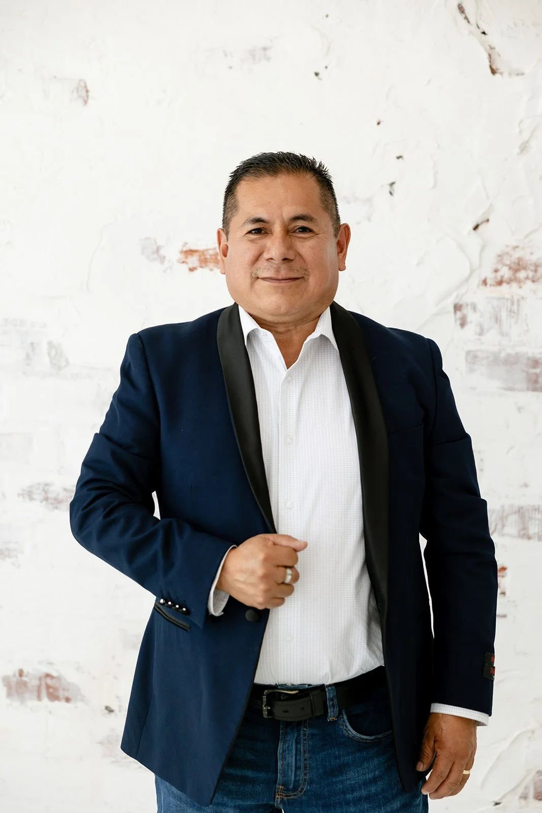 A man in a dark suit jacket, white shirt, and jeans standing in front of a white brick wall.