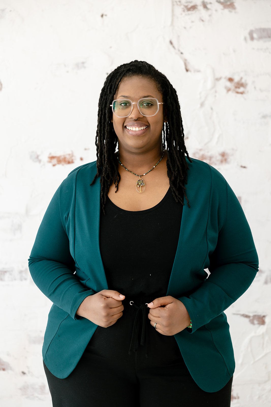 A woman with glasses and dreadlocks, wearing a teal blazer over a black shirt, standing in front of a white brick wall with some red brick showing.