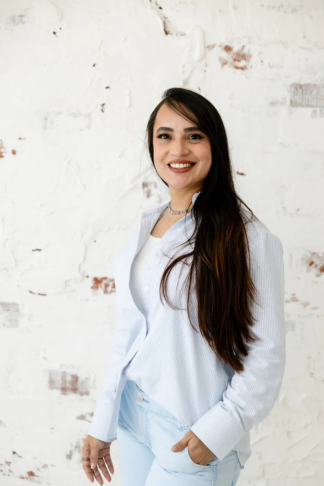 Smiling woman with long dark hair wearing a light blue shirt, white tank top, and light blue jeans, standing in front of a white brick wall.