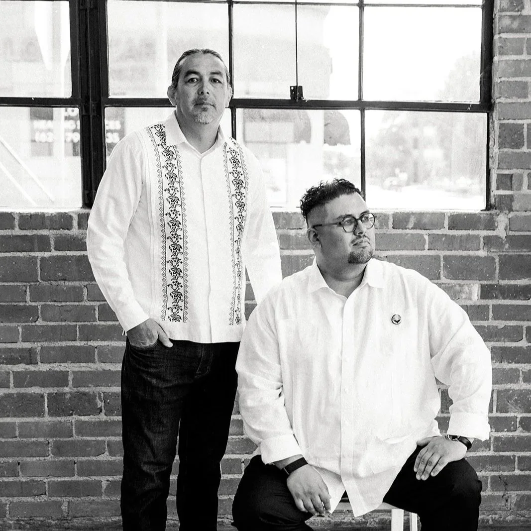 Two men in white shirts posing in front of a brick wall and large windows. One man is standing with one hand in his pocket, and the other is seated, looking to the side.