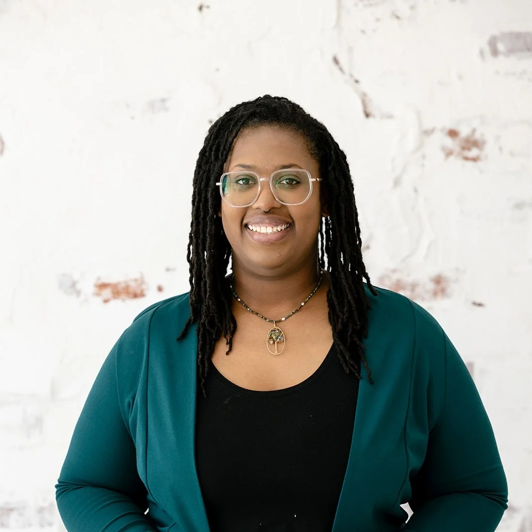 A woman with glasses, black shirt, teal blazer, and a necklace with a pendant. She has dark dreadlocks and is smiling, standing in front of a white brick wall.