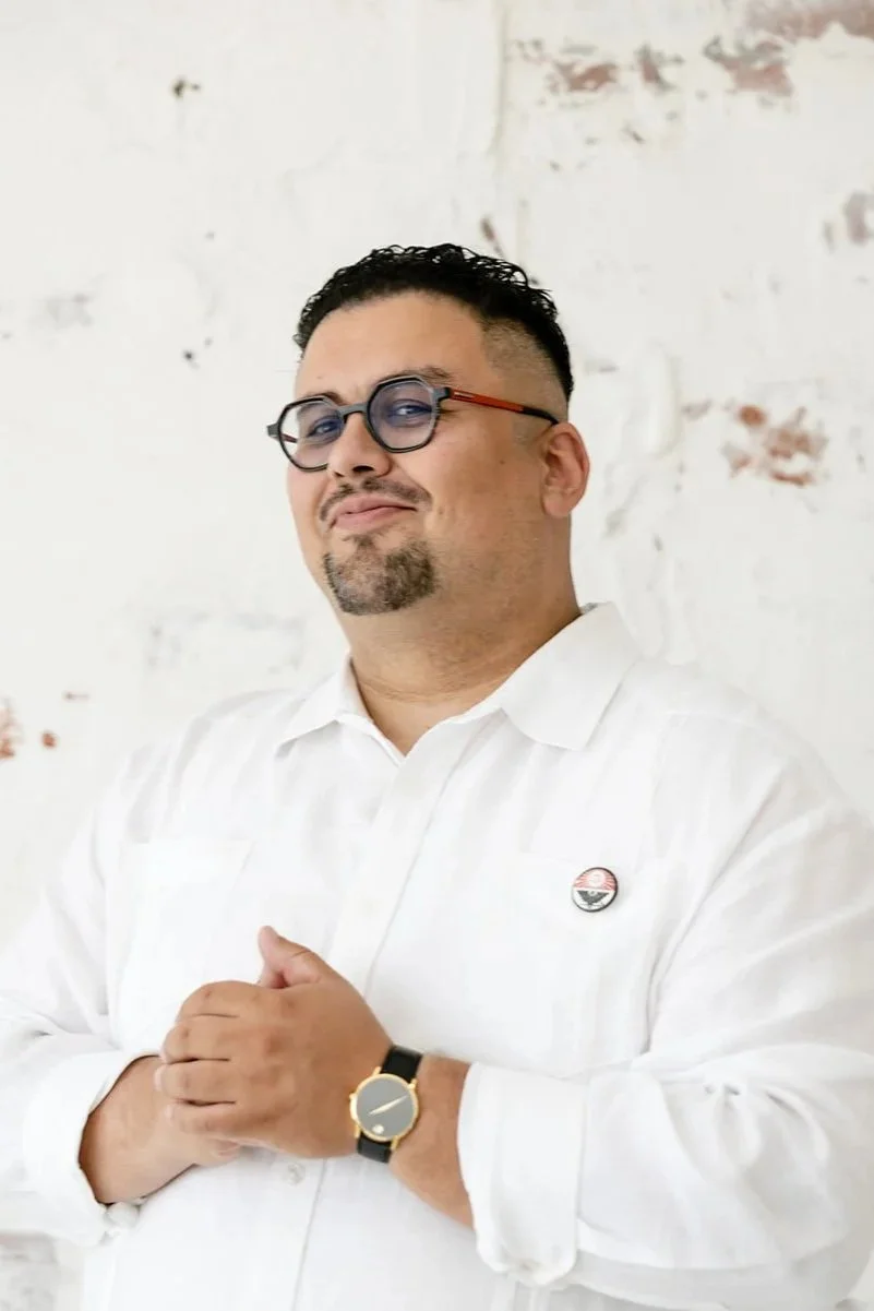 A man wearing glasses and a white dress shirt with a pin on the chest, standing in front of a white textured brick wall.