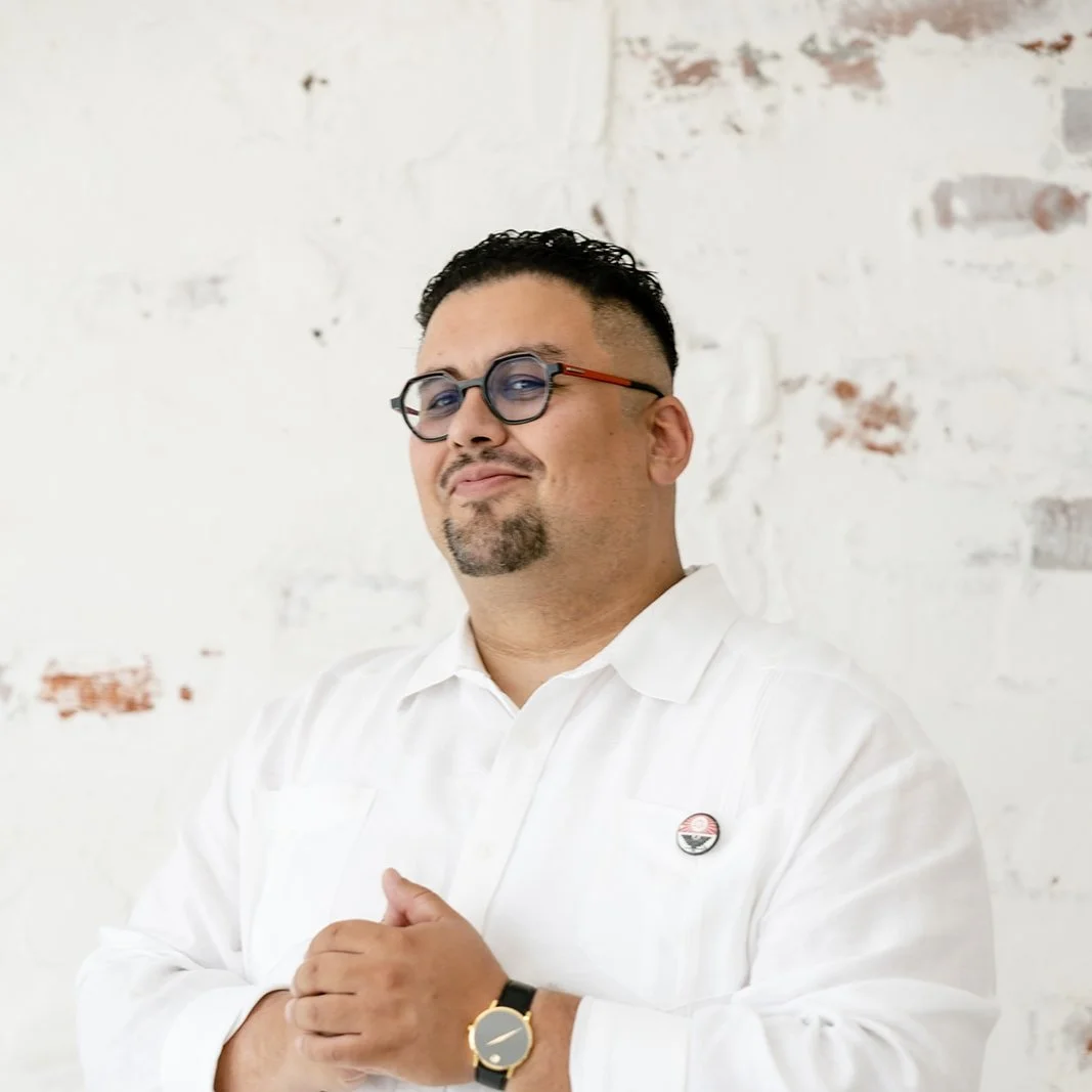 A man with short black hair, glasses, and a goatee, wearing a white shirt with a small pin, a black watch, stands in front of a white brick wall with some peeling paint.