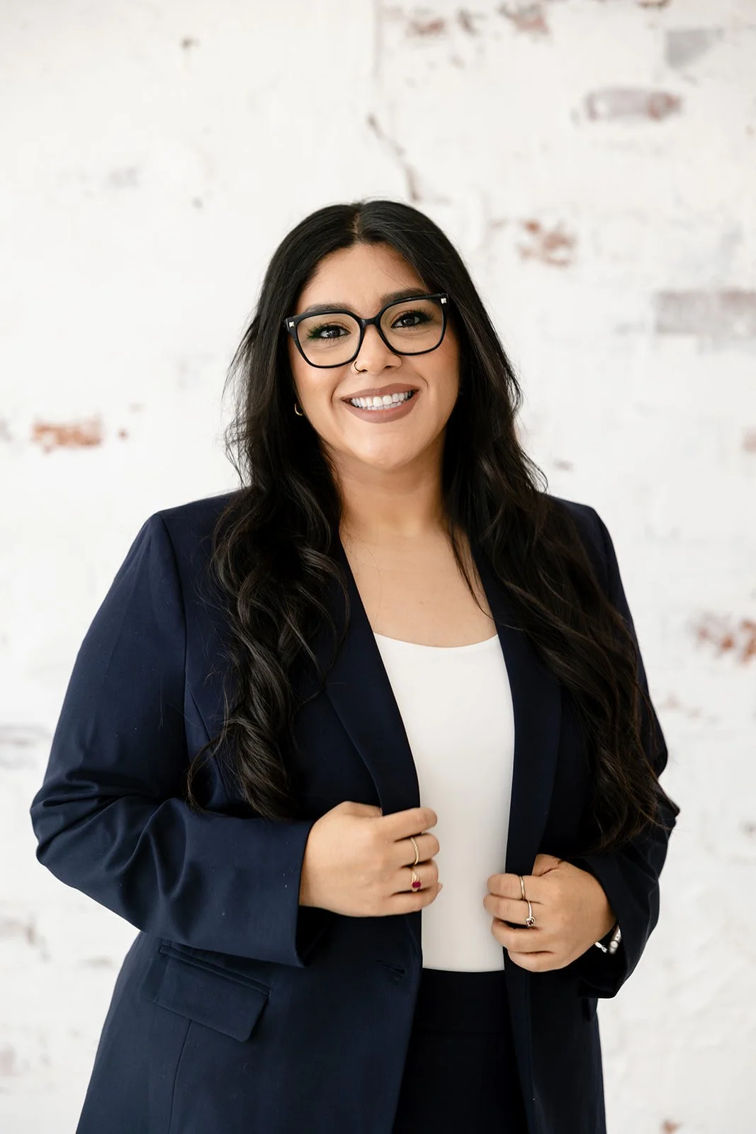 A woman wearing glasses and a navy blazer, smiling, with long dark hair and a white top, standing against a white brick wall.