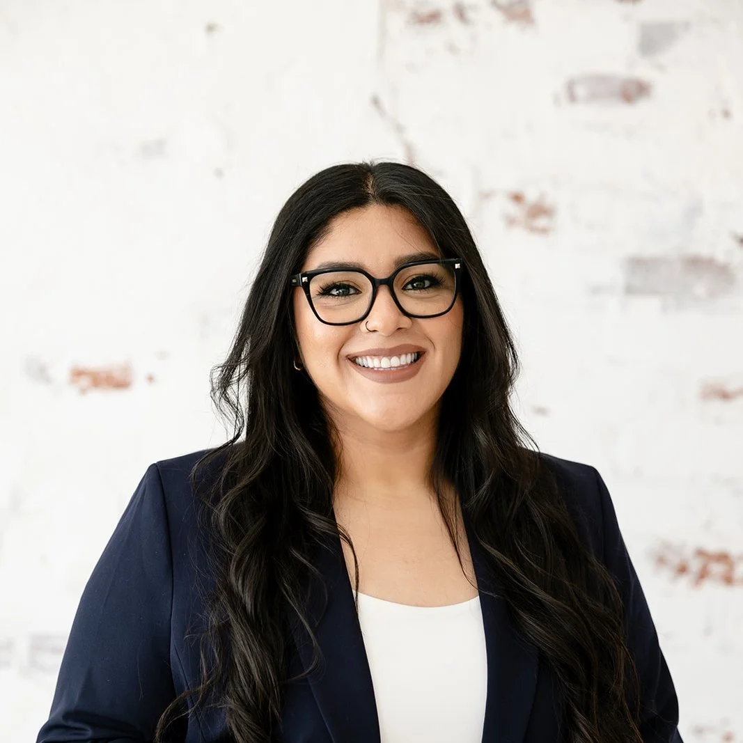 Portrait of a woman with long dark hair, glasses, and a friendly smile, wearing a navy blazer and a white top, standing against a white brick wall.