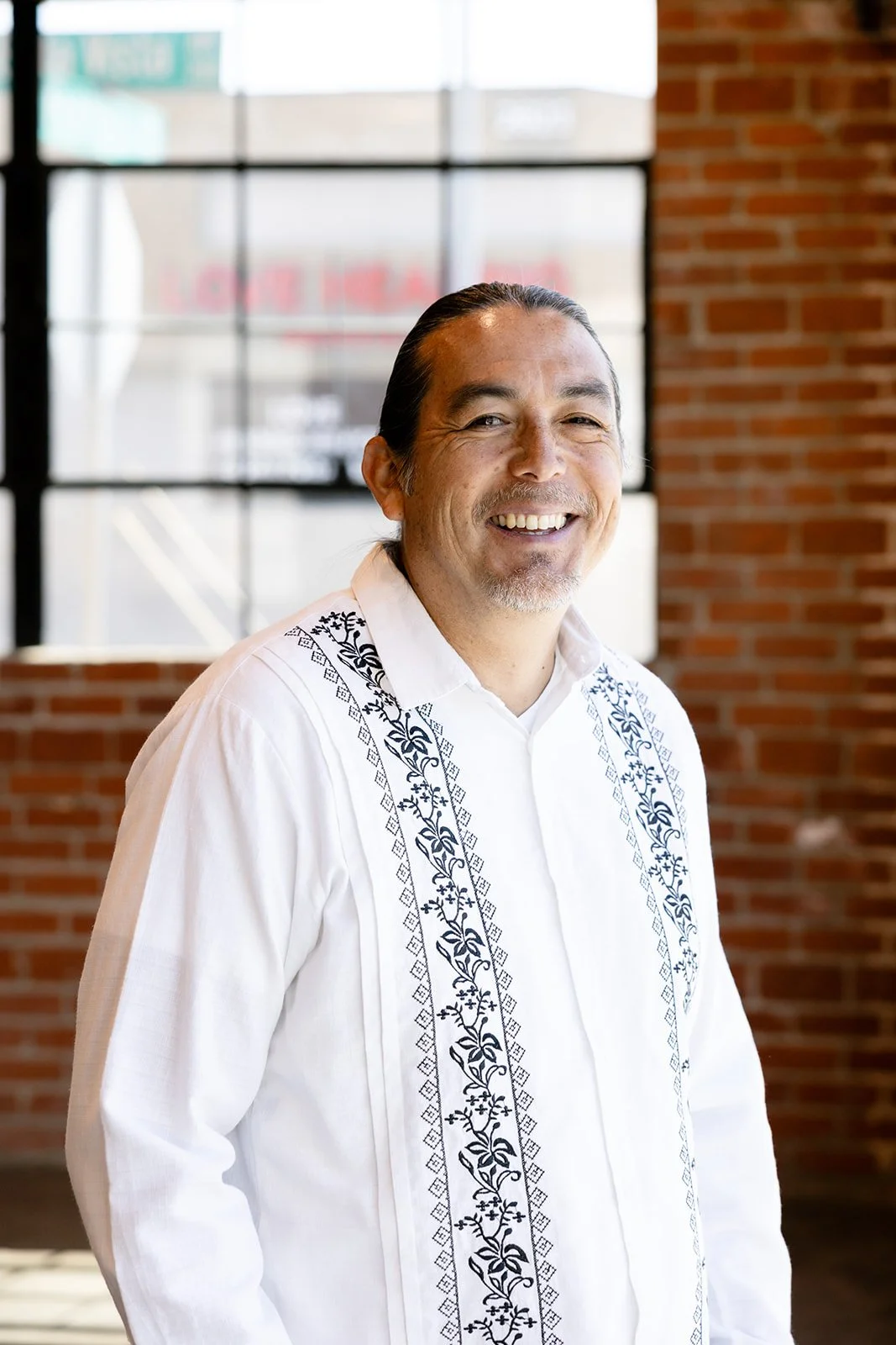 A smiling man in a white embroidered shirt standing indoors near a brick wall and large windows.
