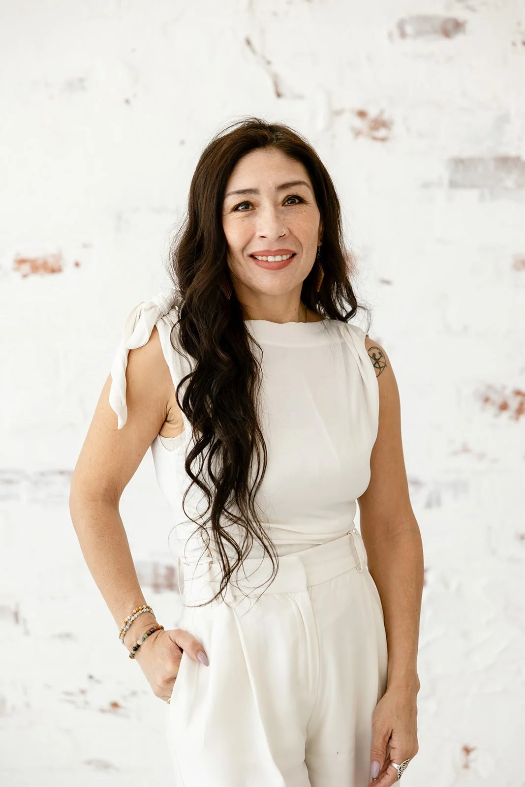 A woman with long dark wavy hair, wearing a white sleeveless top with tied shoulder straps, standing against a white brick wall, smiling with her right hand in her pocket.