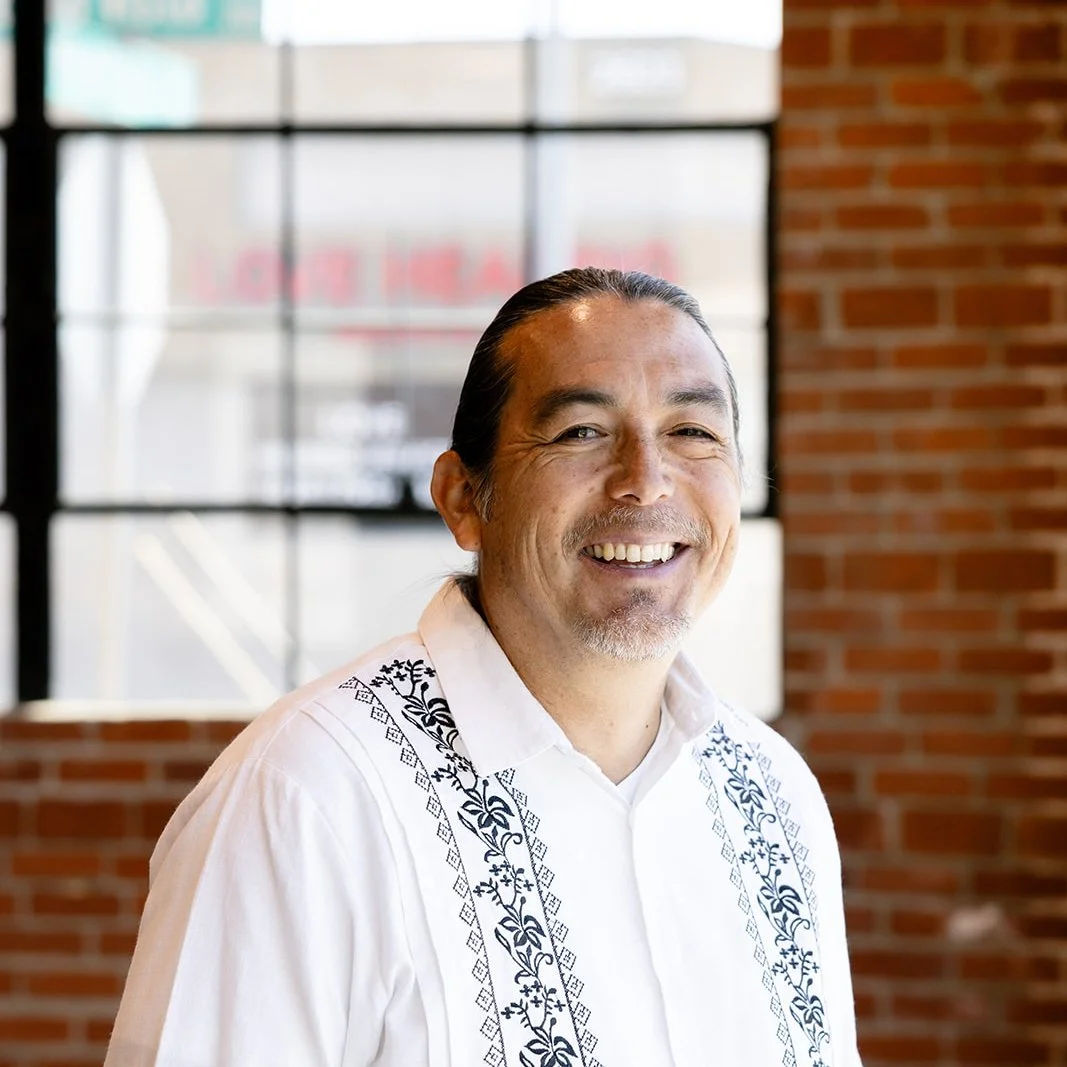 A smiling man with long dark hair tied back, wearing a white shirt with black embroidered designs, standing indoors with a brick wall and large window in the background.