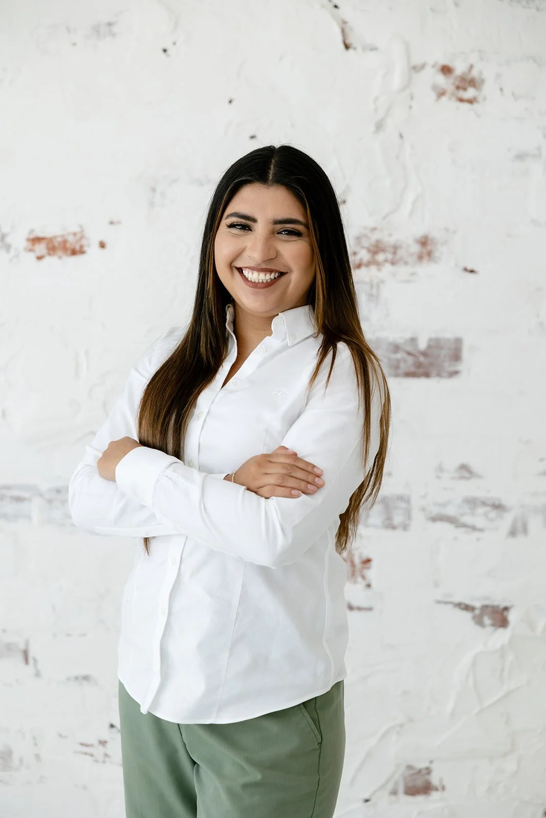 A woman with long dark hair smiling with arms crossed, wearing a white shirt and olive-green pants, standing against a white brick wall.