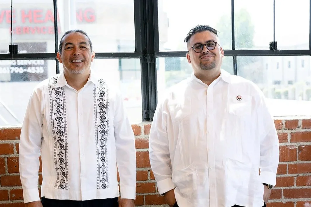 Two men standing indoors in front of a large window with a brick wall behind them. The man on the left has long hair, a beard, and wears a white shirt with black embroidered patterns. The man on the right has short dark hair, glasses, a goatee, and wears a plain white shirt with a small pin on the pocket.