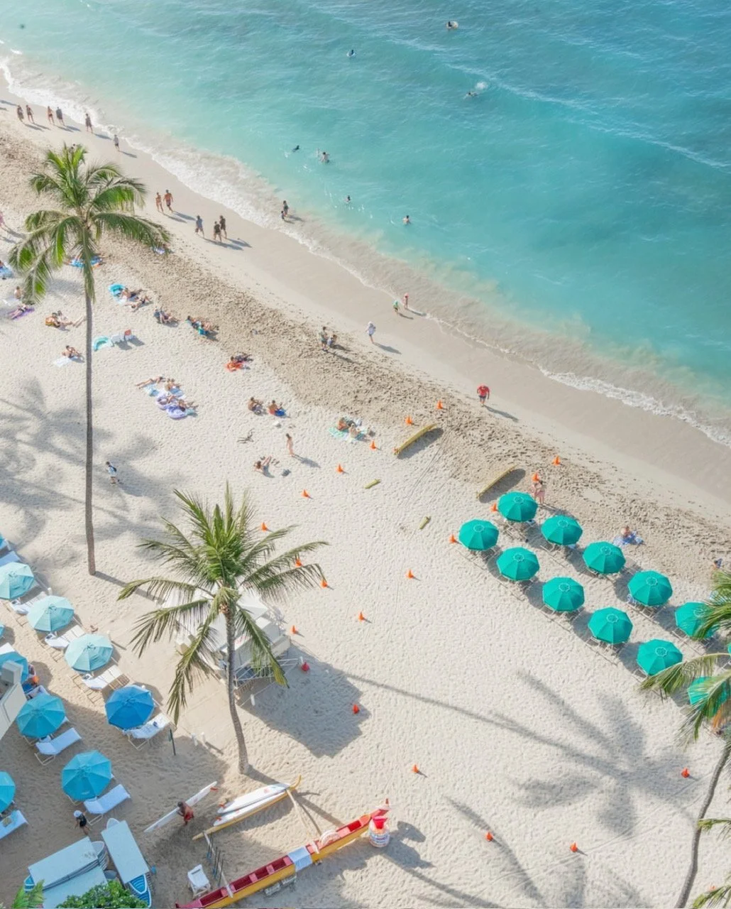 Crowded beach with beach chairs and umbrellas, palm trees, people swimming and sunbathing, crews and beach patrol markers, and shallow turquoise water.