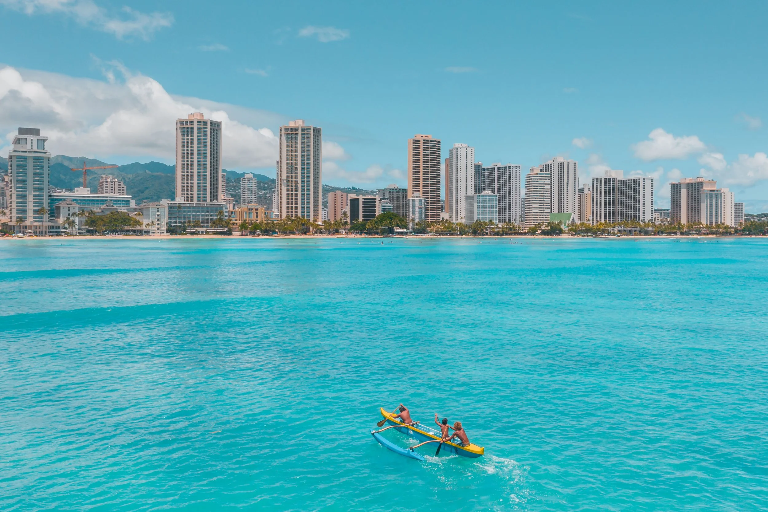 Three people kayaking in turquoise water with a city skyline and mountains in the background.
