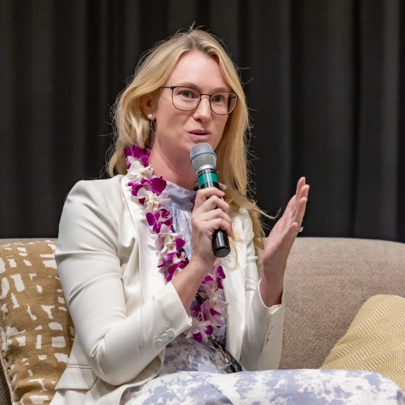 A woman with blonde hair, glasses, and pearl earrings speaking into a microphone while sitting on a beige couch, wearing a white blazer and a purple and white lei.