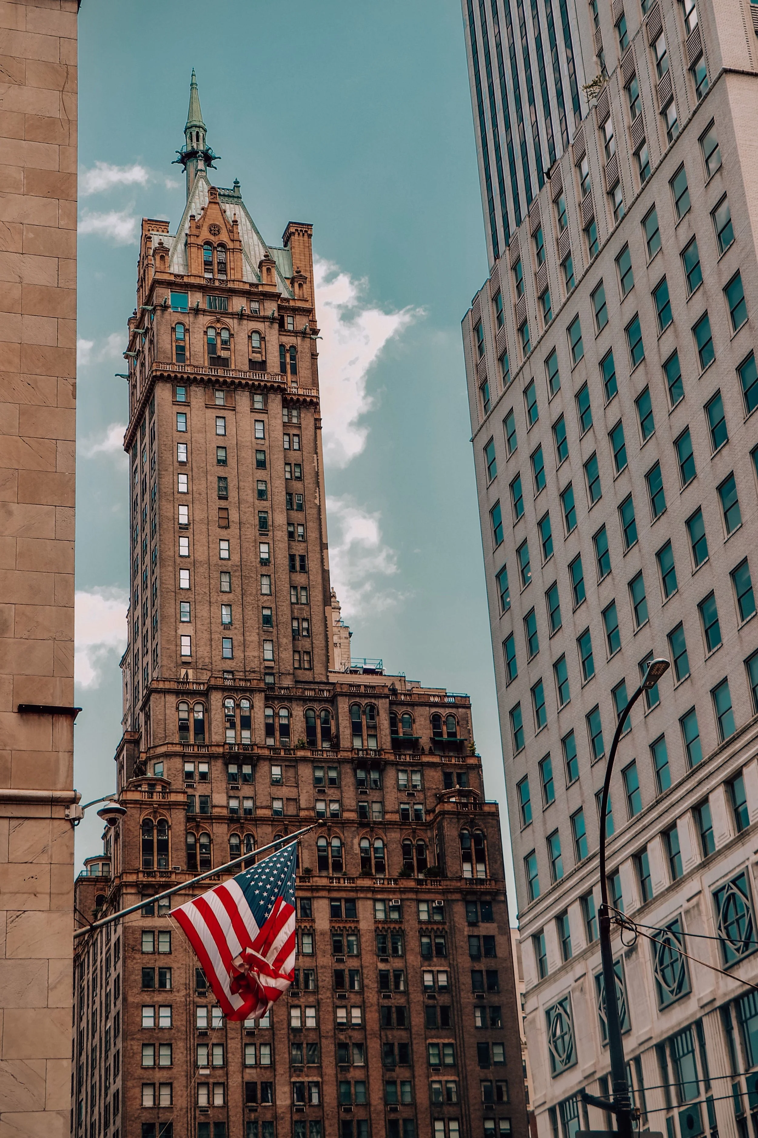 A city street with tall skyscrapers and iconic buildings, including a historic tower and modern glass buildings. An American flag is displayed in the foreground, with a street lamp visible nearby.