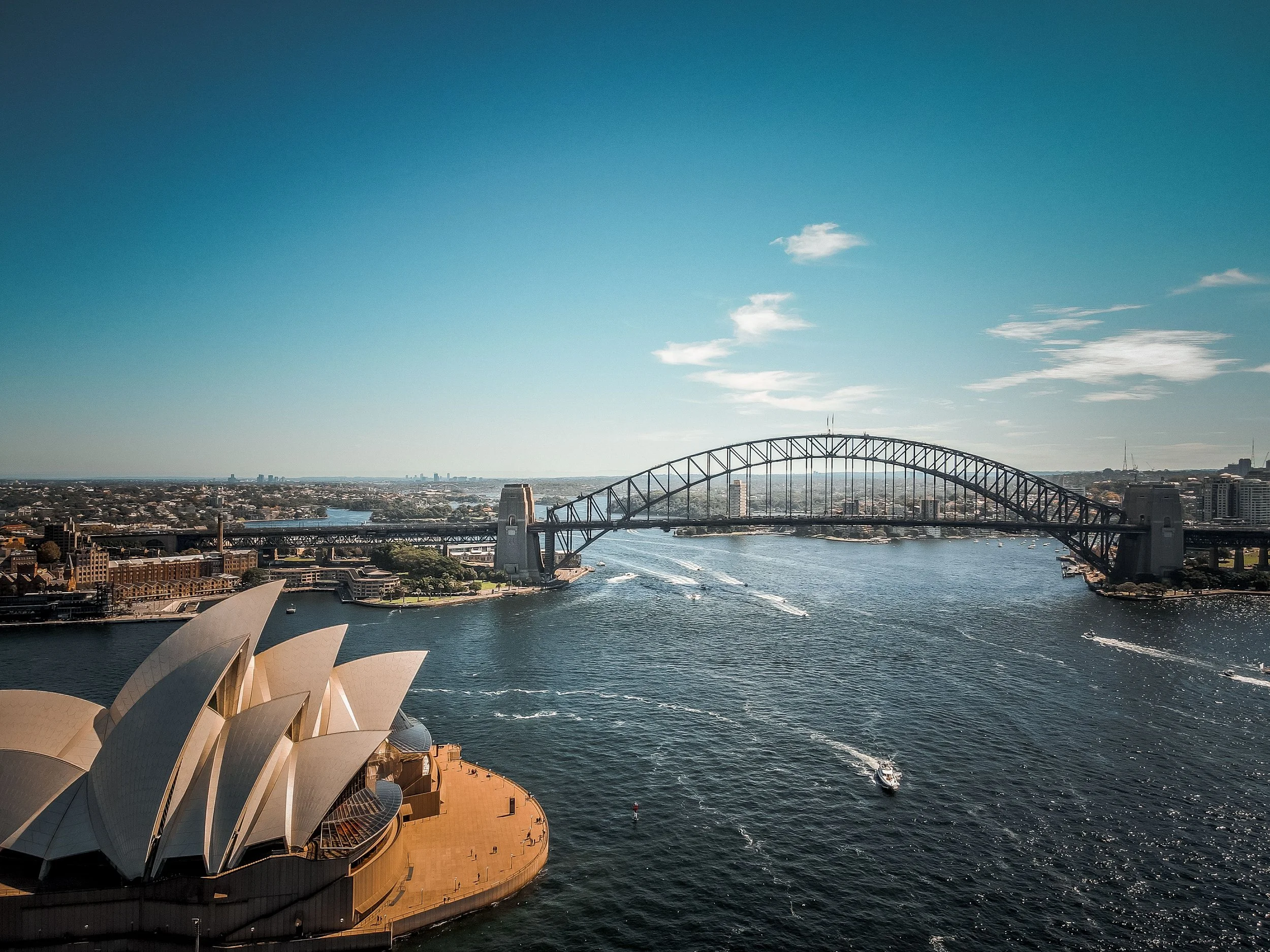 Sydney Opera House in the foreground with the Sydney Harbour Bridge in the background, boats on the water, and a city skyline under a partly cloudy blue sky.