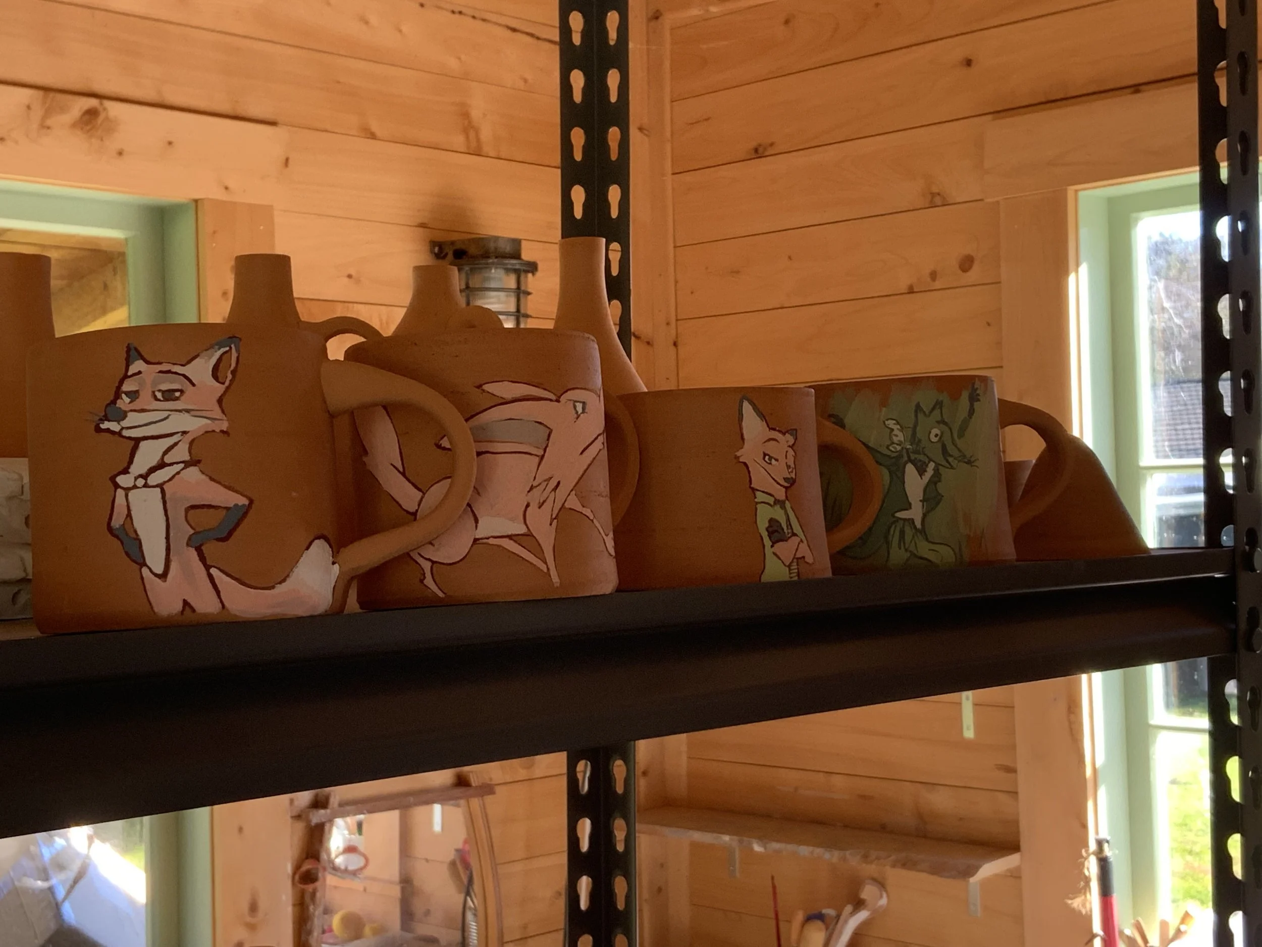 Shelves holding mugs in progress with decorations, inside a room with wooden walls and windows.