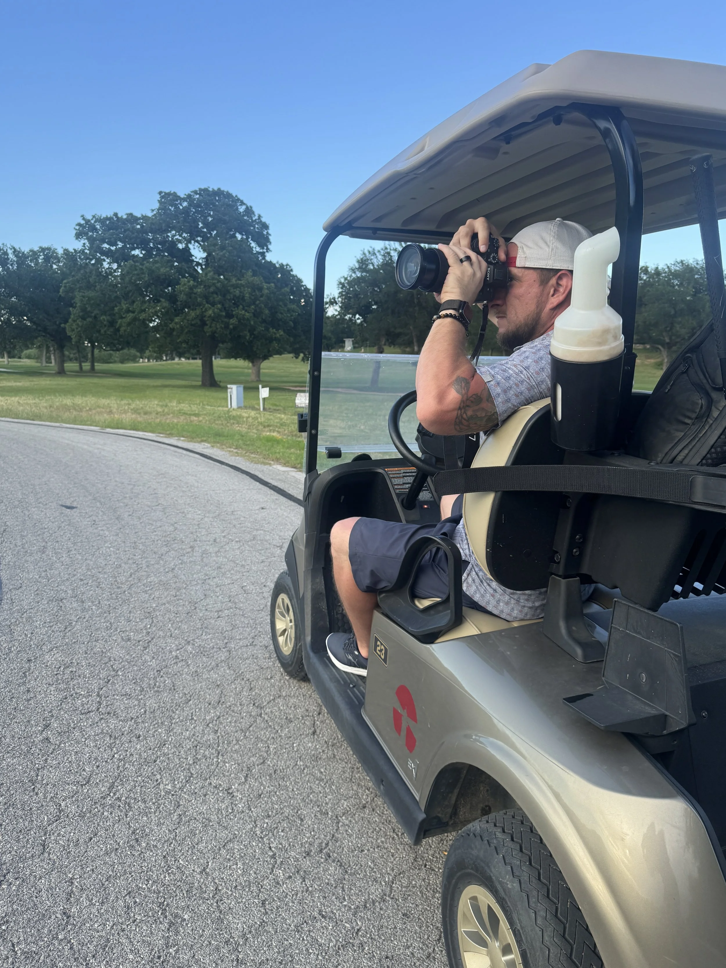 A man sitting in a golf cart, taking pictures with a camera, wearing a baseball cap, a watch, a bracelet, and casual clothing, parked on a paved path with trees and a clear blue sky in the background.