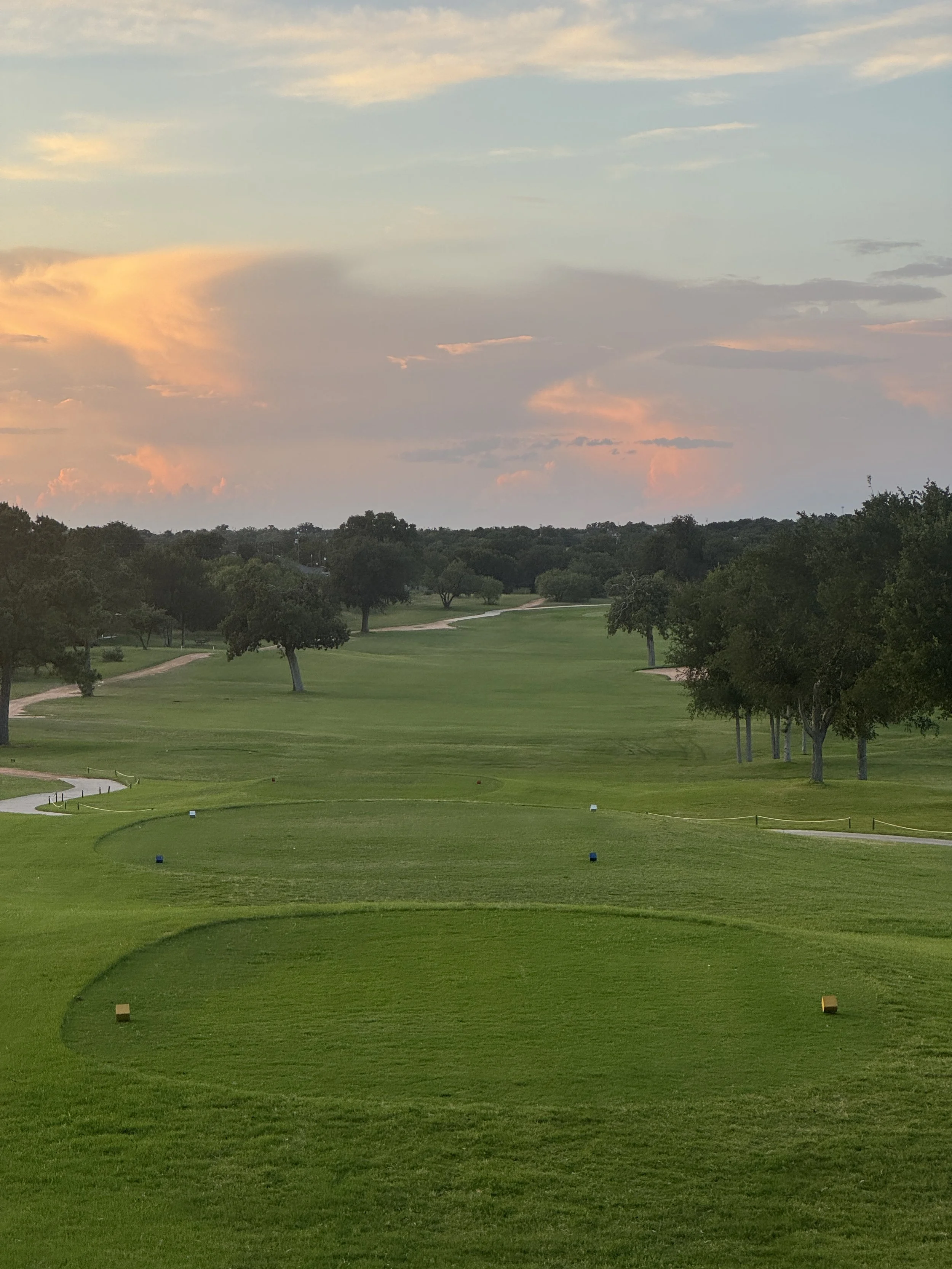 A golf course at sunset with lush green grass, trees, and a winding path, under a partly cloudy sky with soft pink and blue clouds.