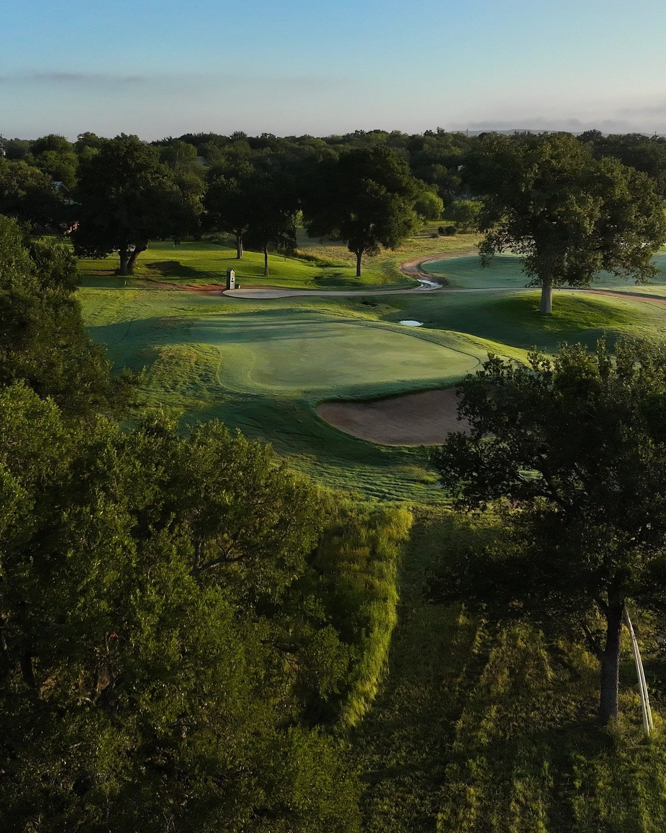 A scenic view of a golf course with green grass, trees, a small water hazard, a sand bunker, and a walking path, taken during late afternoon or early evening.
