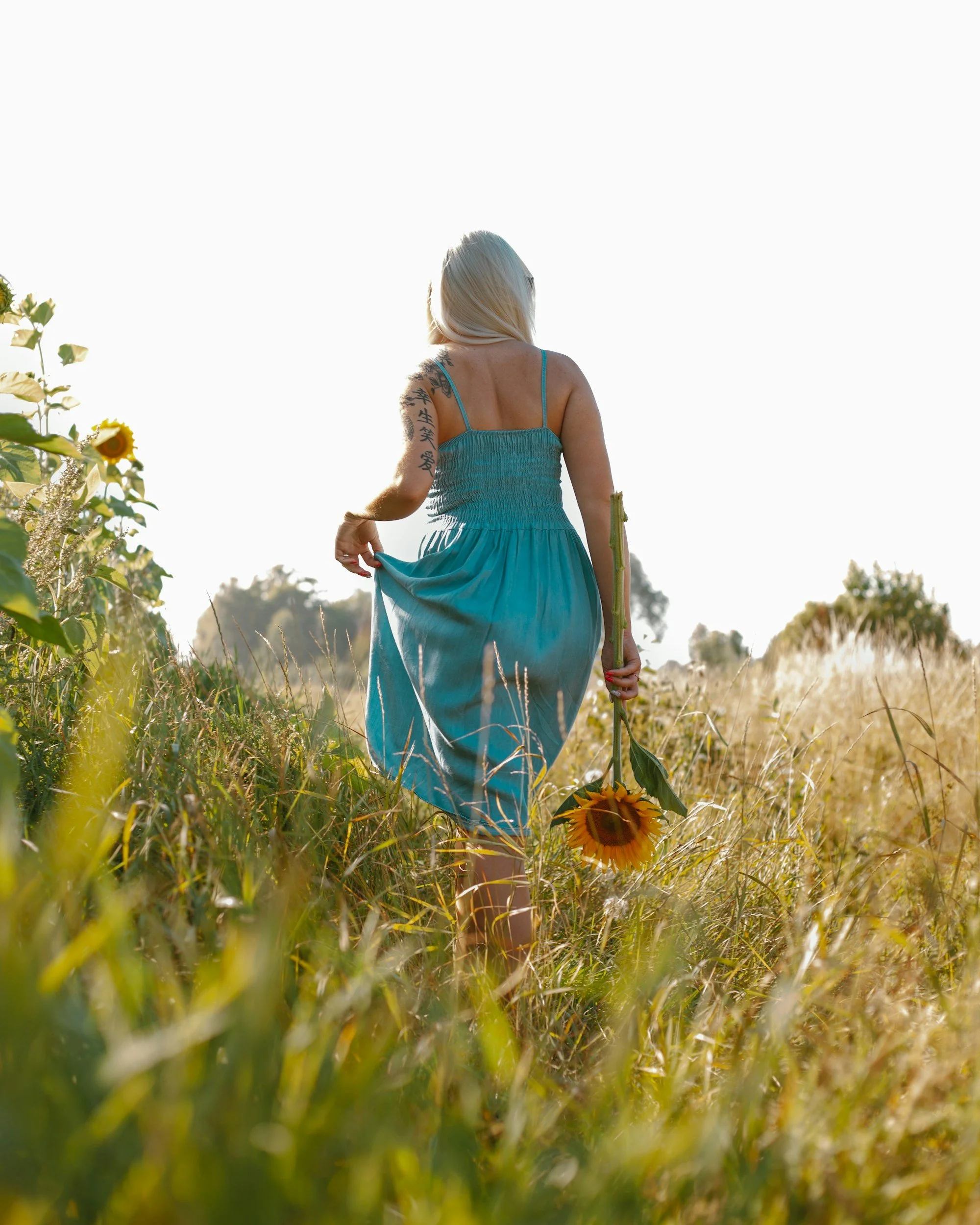 A woman with long blonde hair, wearing a blue sundress, walking through a field of tall grass and sunflower plants during sunset or sunrise, holding a sunflower in her right hand.