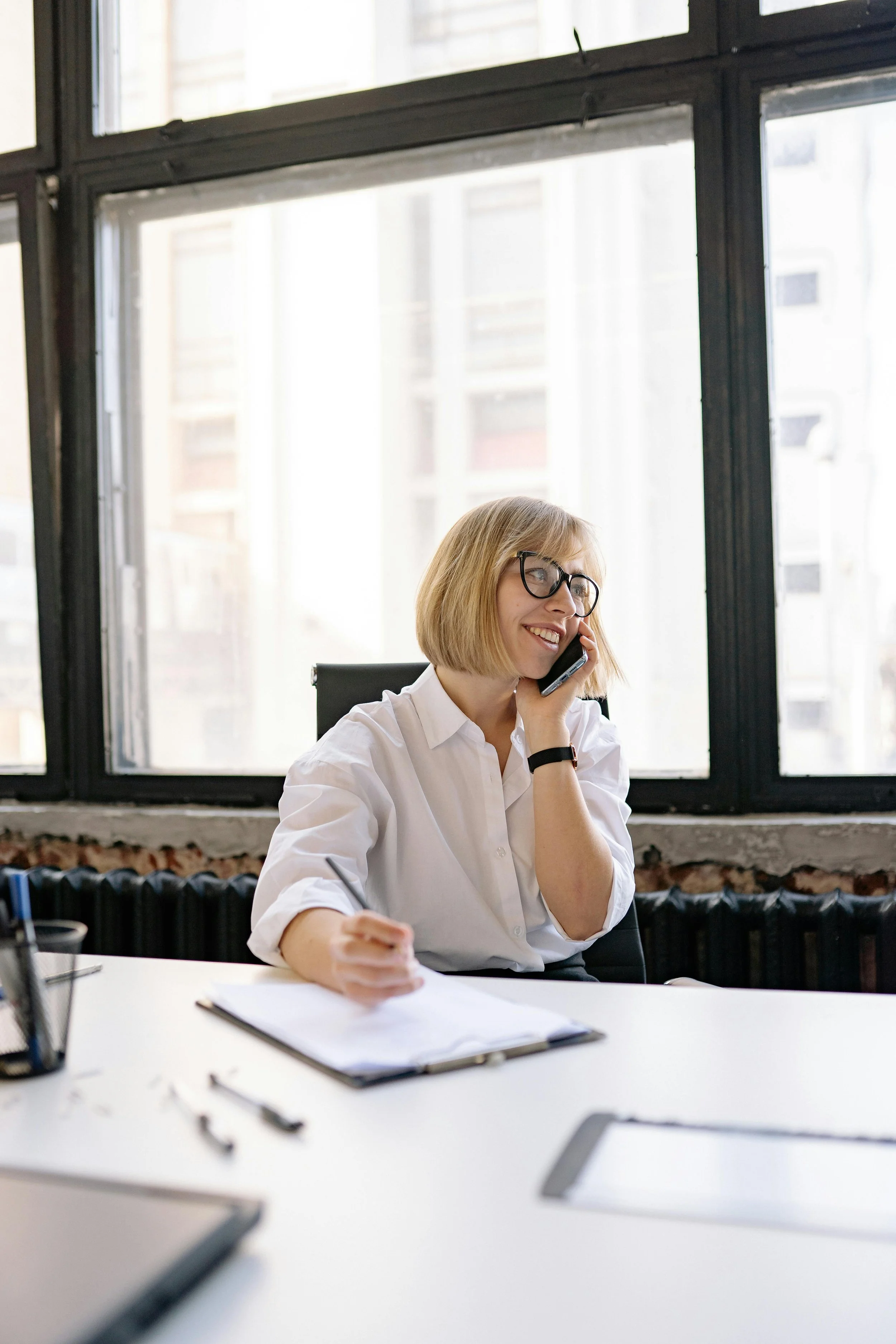 A woman with blonde hair and glasses sitting at a desk in front of large windows, talking on a mobile phone, with papers and stationery on the desk.