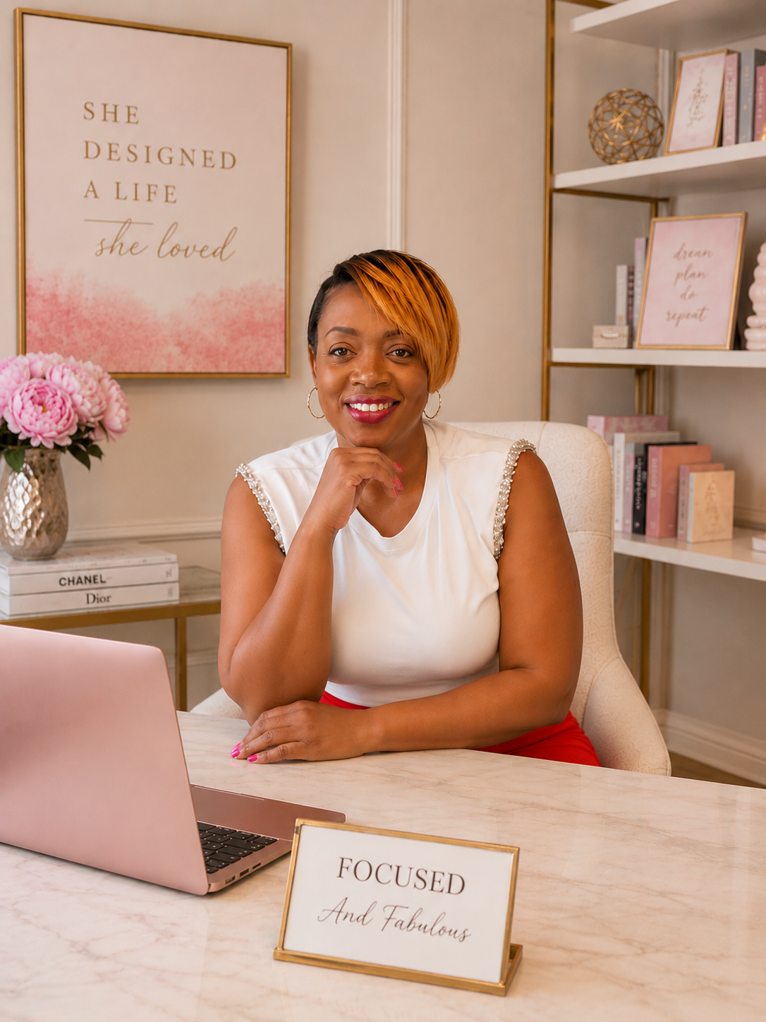 A woman with short hair and hoop earrings sitting at a desk with a pink laptop, smiling, in a decorated office with inspirational posters and books. A sign on the desk reads "FOCUSED And Fabulous."