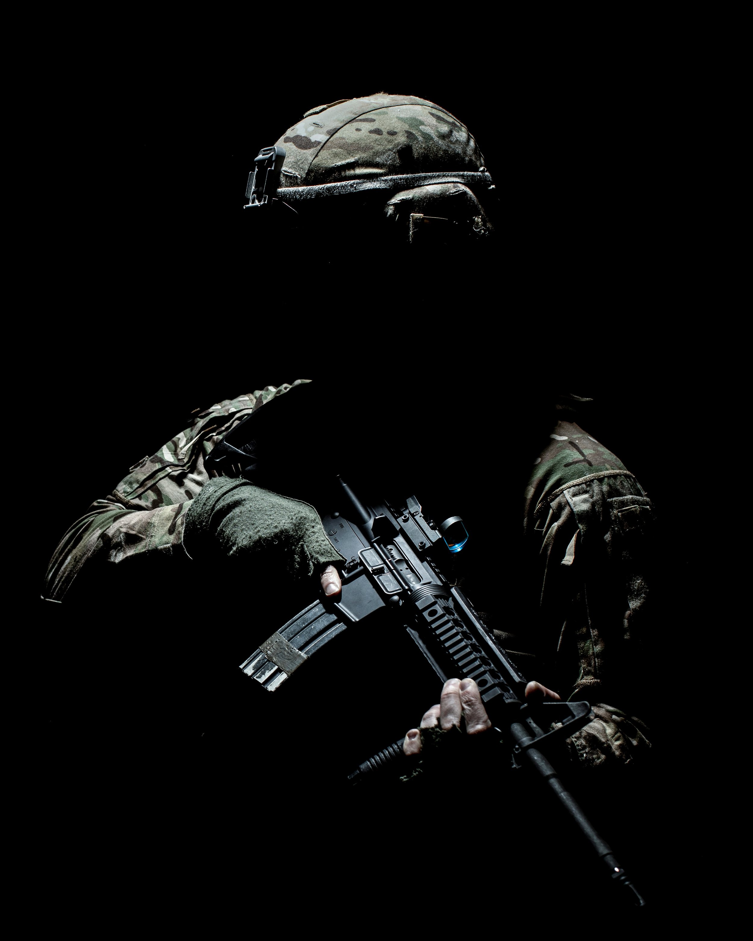 A soldier wearing camouflage uniform and helmet, holding a rifle, against a black background.
