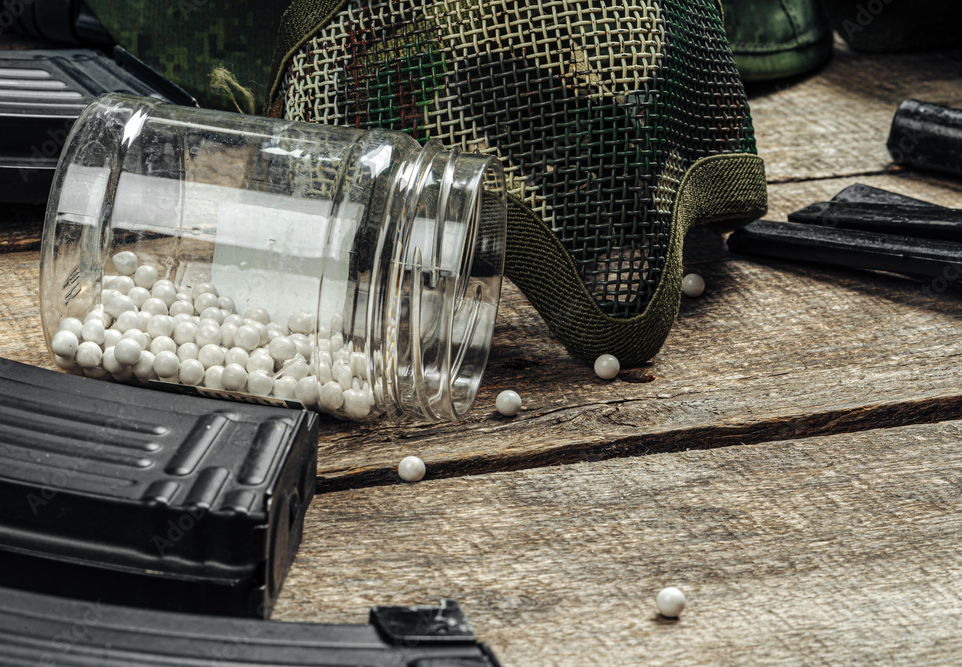 A clear plastic container of white paintballs on a wooden surface with some paintballs spilled out, near a camouflage paintball mask and black paintball magazines.