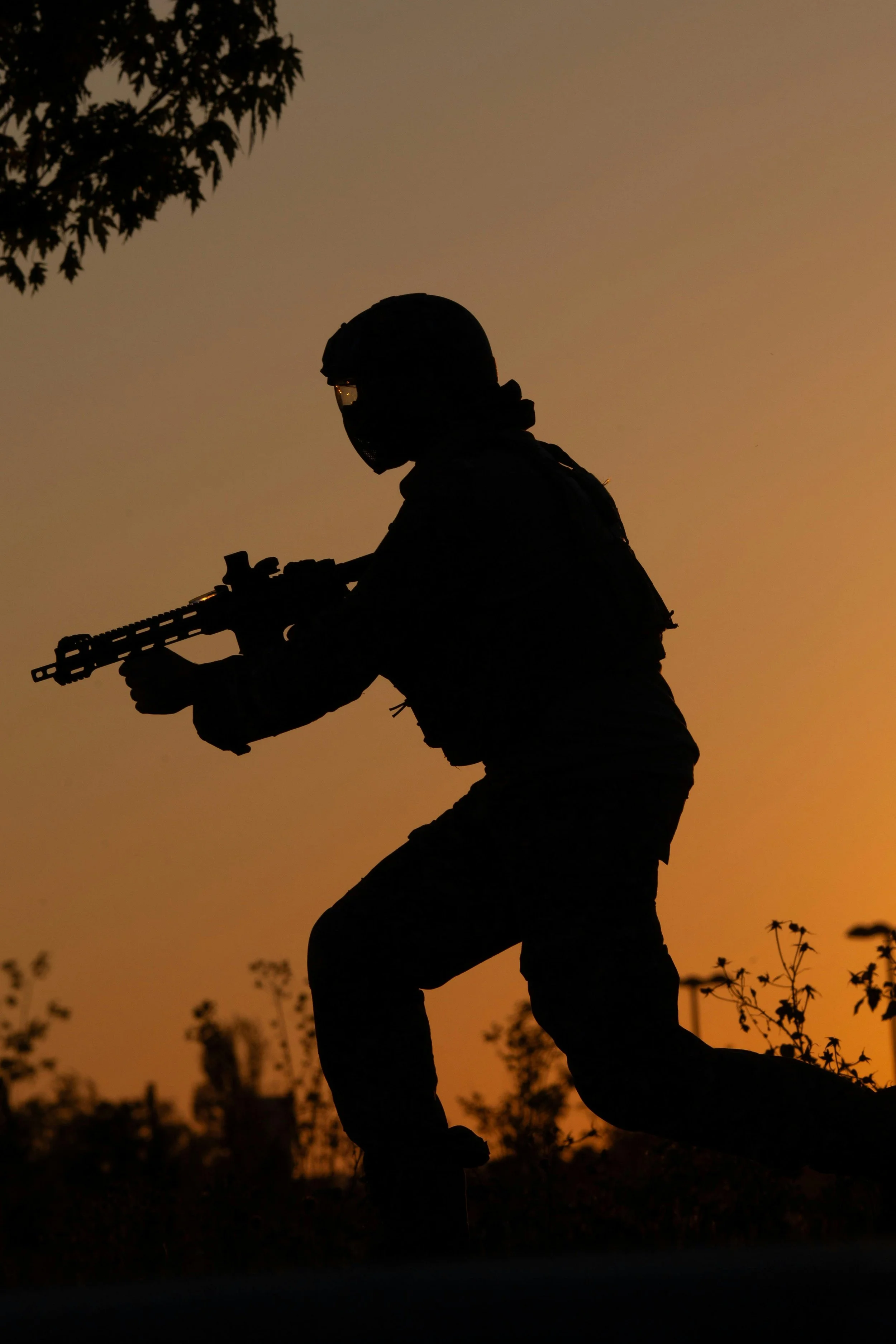 Silhouette of a soldier holding a rifle, in a crouched position, with a sunset or sunrise sky in the background and trees in the foreground.