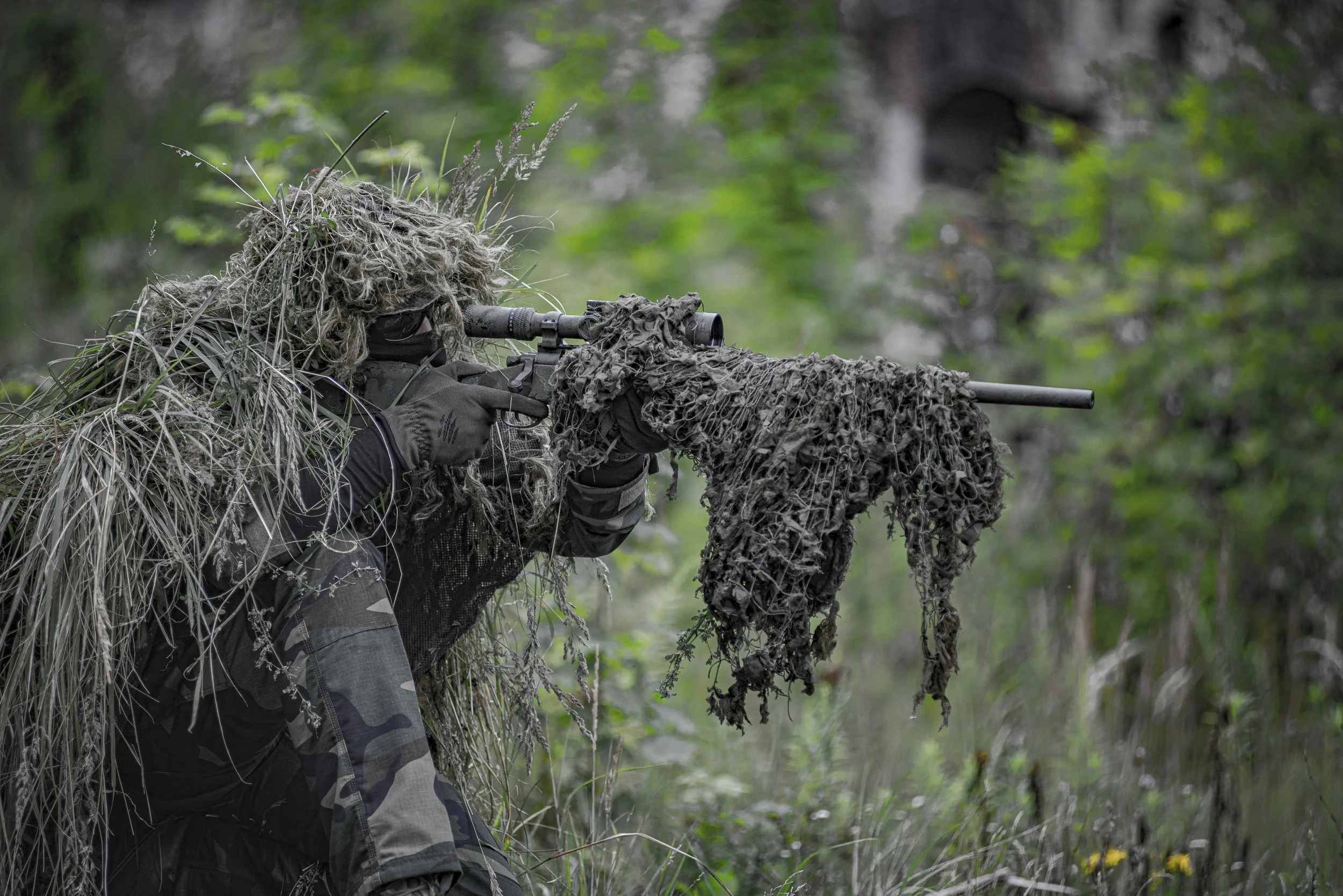 A soldier dressed in camouflage gear aiming through a sniper scope in a forested area.