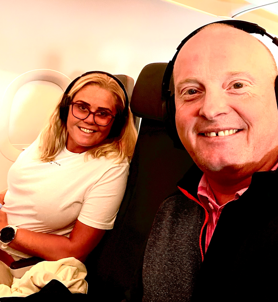 A woman and a man sitting next to each other on an airplane, both wearing headphones and smiling at the camera.