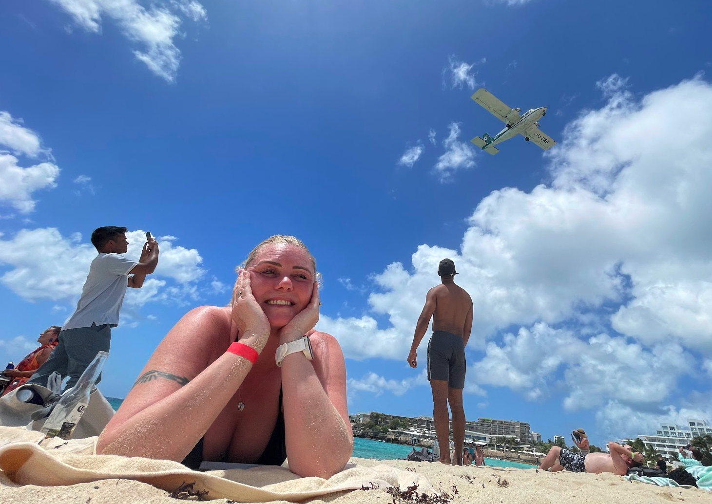 Beach scene with a smiling woman lying on the sand at the beach in Saint Martin, a small plane flying overhead in a partly cloudy sky, and people relaxing and taking photos in the background.