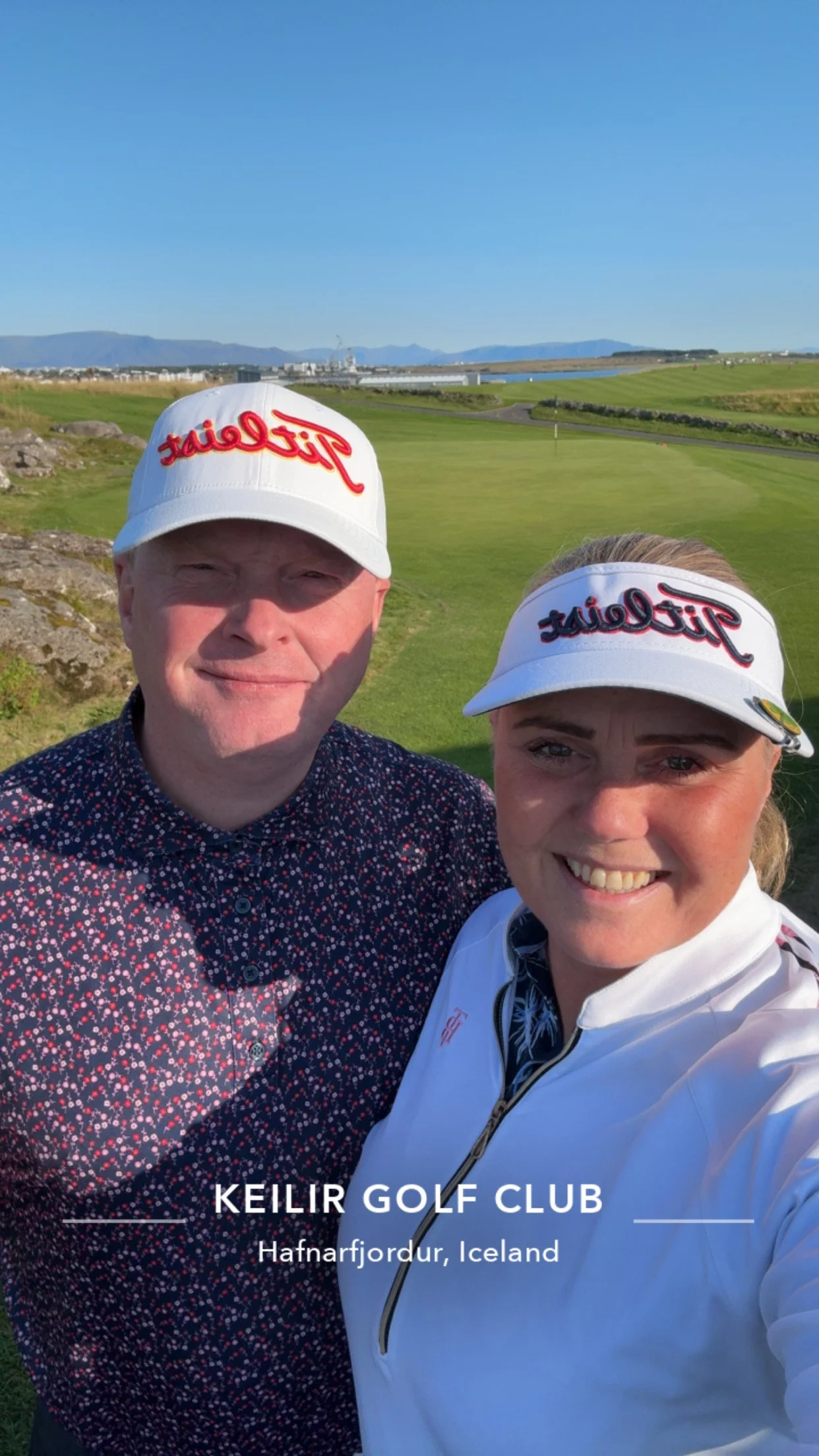 Two people taking a selfie at Keilir Golf Club in Hafnarfjörður, Iceland, on a sunny day with a clear blue sky, green golf course, and mountains in the background, both wearing white caps with red and black logos.