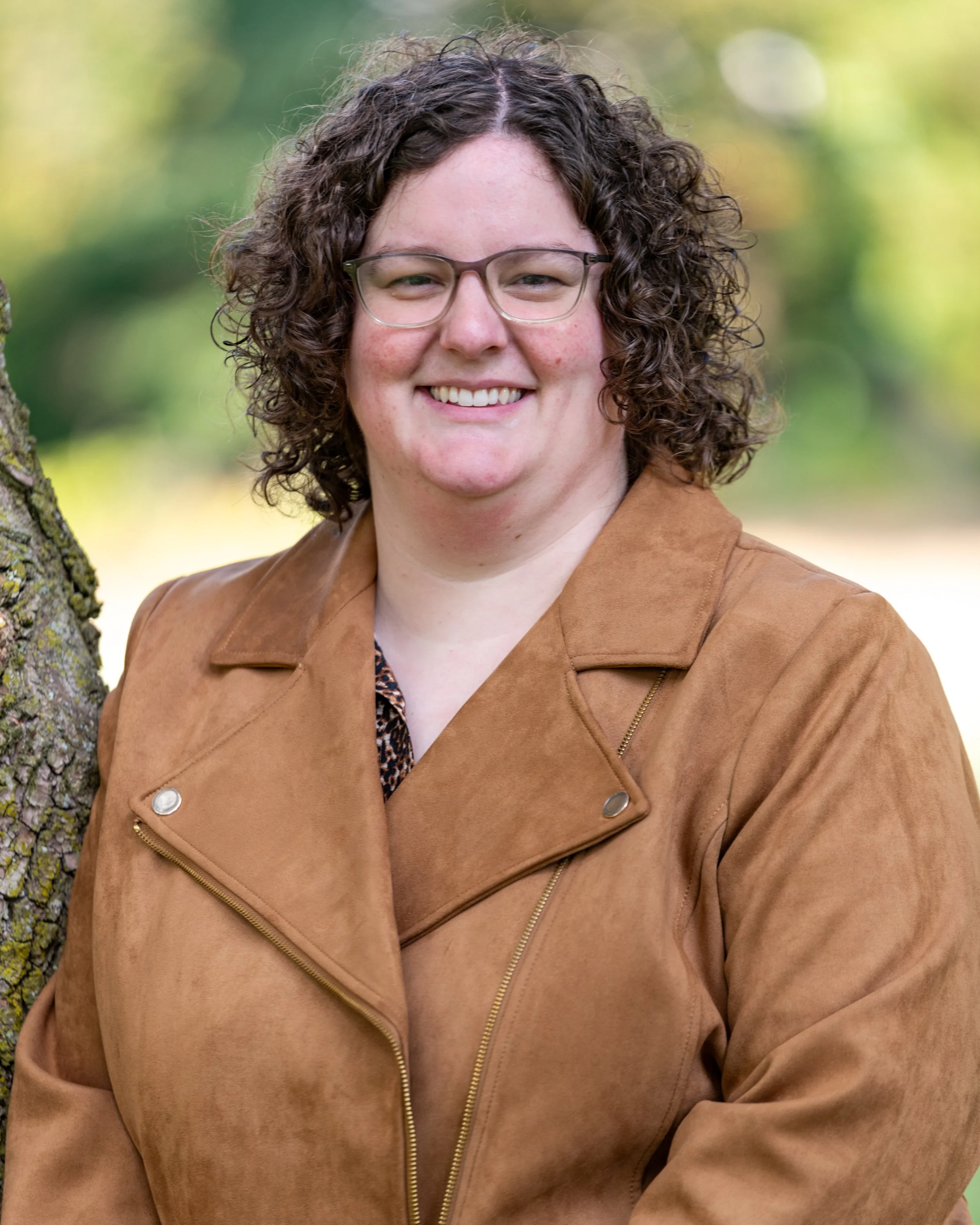 A woman with curly brown hair, glasses, and a tan jacket smiling outdoors with a blurred green background.