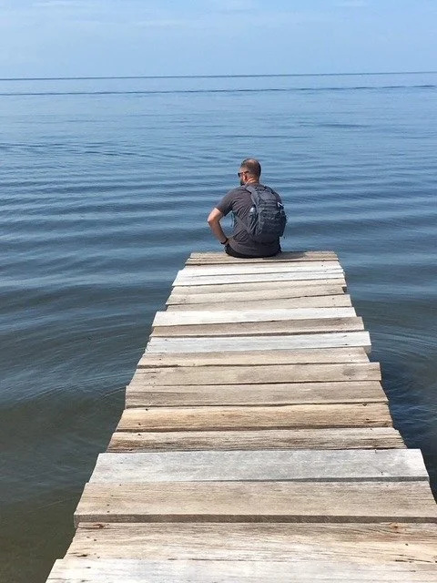 Man sitting on a wooden dock by a calm lake, with a backpack, looking at the water.